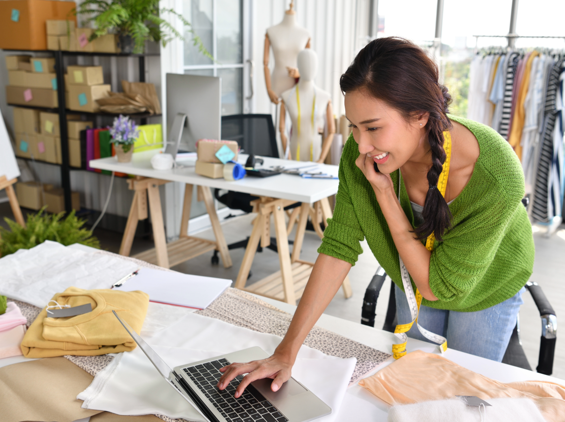 A woman in a green sweater with a measuring tape around her neck working on a sewing project at a table covered with fabric and a laptop in a craft room. There are dress forms mannequins, a rack of clothing, and shelves with boxes and supplies in the background.