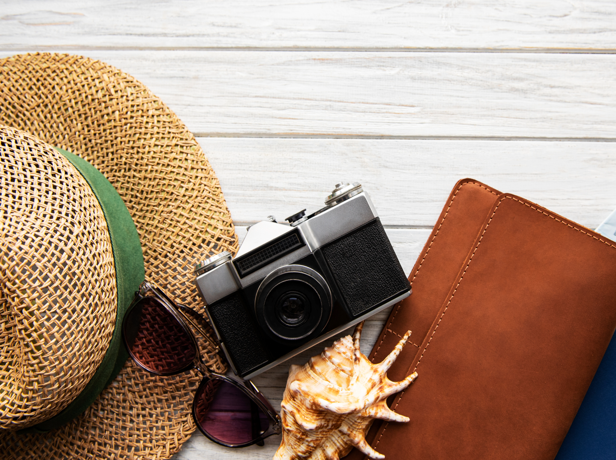 A flat lay of summer vacation items, including a straw hat, sunglasses, an old camera, seashells, and brown leather journal.