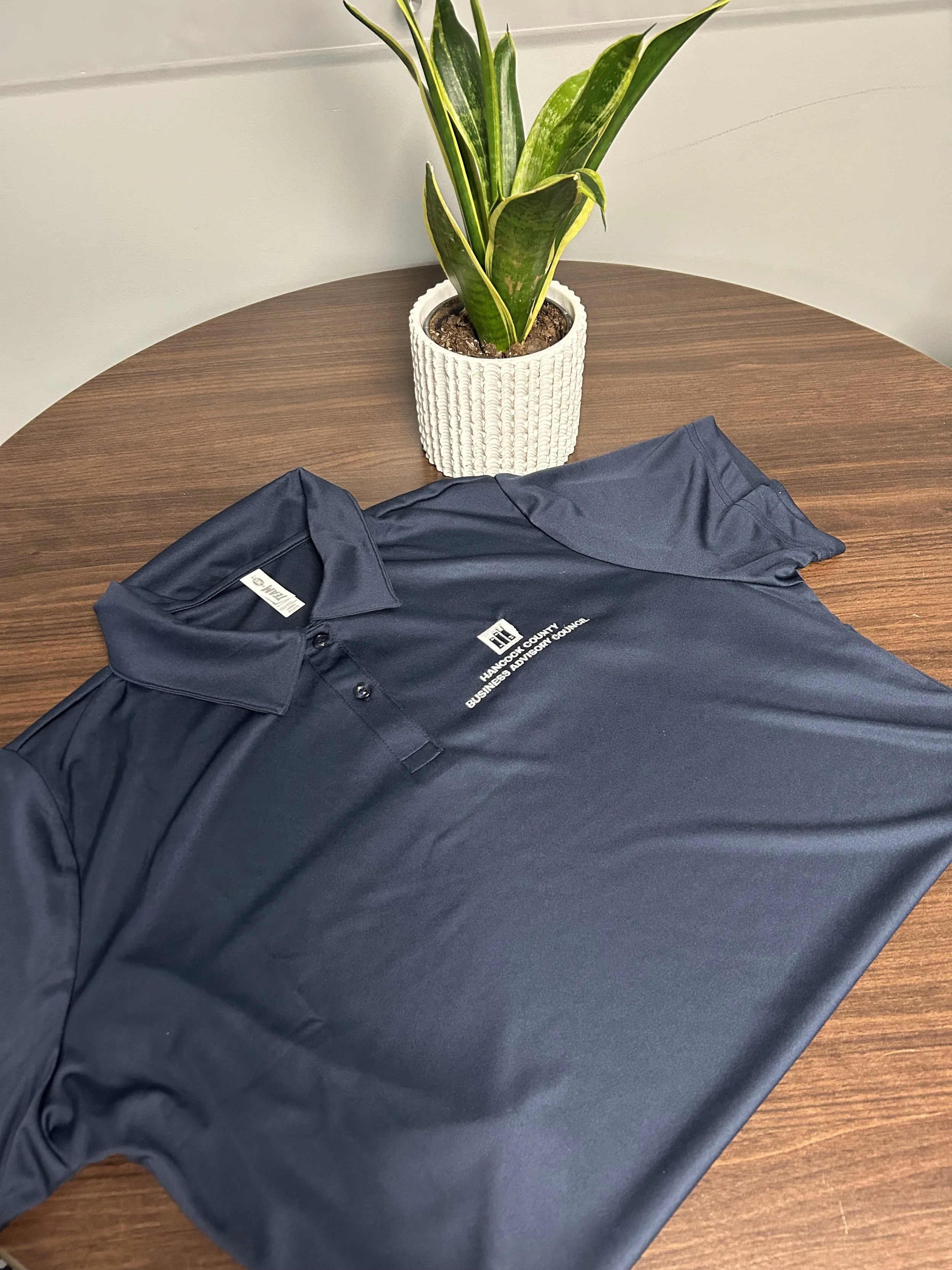 A dark blue polo shirt with printed text 'Hancock County Business & Advisory Council' lies on a wooden table.