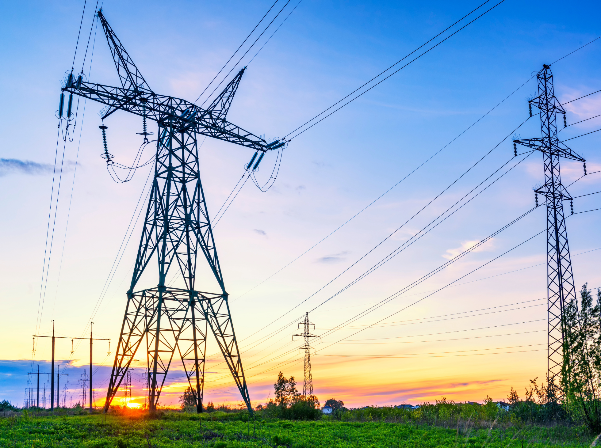 Electric transmission towers standing in a field at sunset, with multiple power lines extending into the distance.