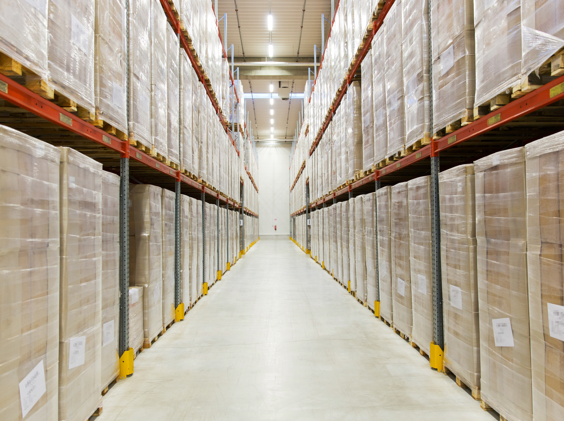 Warehouse aisle with tall shelving units filled with stacked boxes wrapped in plastic.