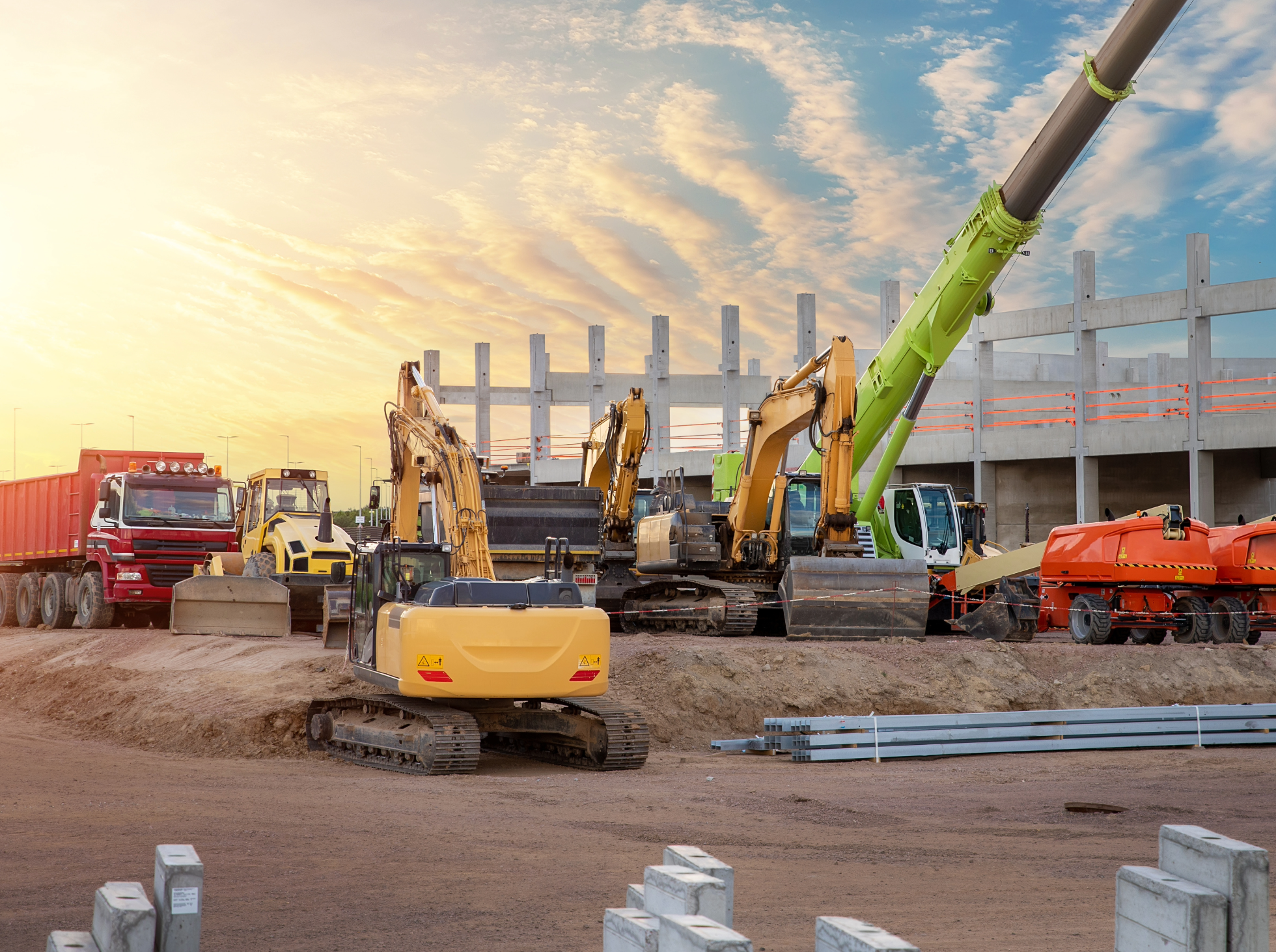 Construction site with multiple heavy machinery machines including excavators, a crane, a truck, and construction materials with a partially built structure.