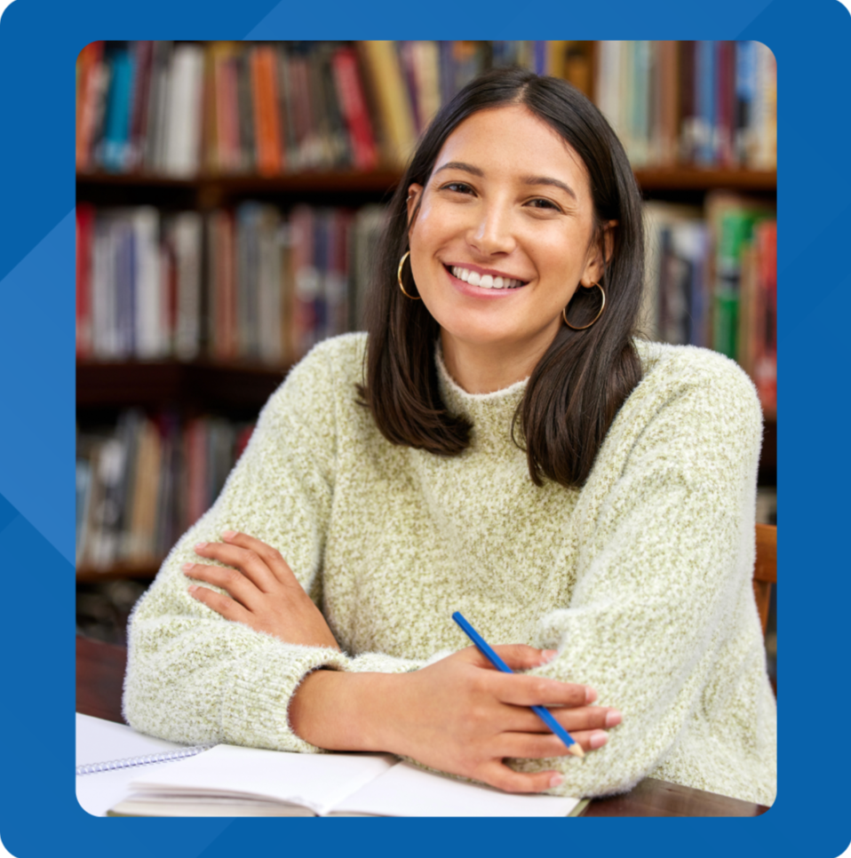 A young woman with dark hair and gold hoop earrings smiling at the camera, sitting at a table with a notebook and a blue pen, in front of a bookshelf filled with books.