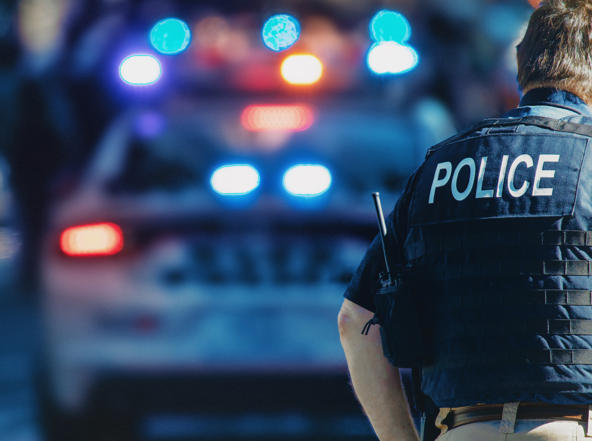 A police officer wearing a vest with the word 'POLICE' on the back, standing at night with police cars with flashing lights in the background.