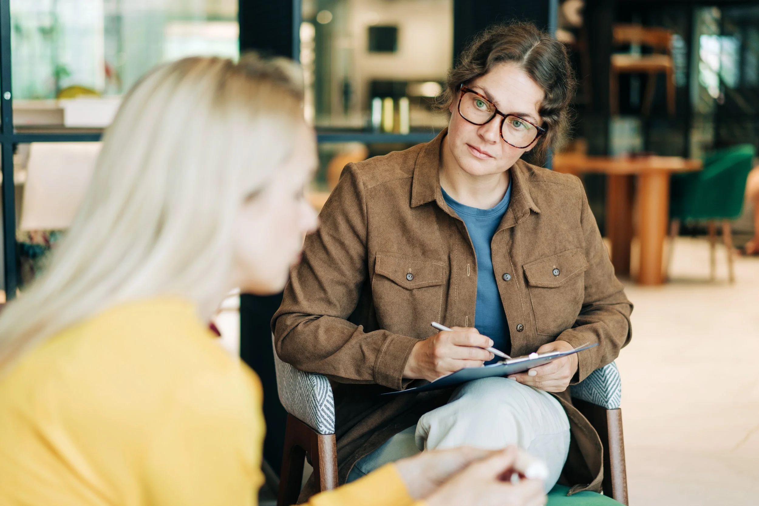 A woman sitting at a table, writing in a notebook, engaged in conversation with another woman.