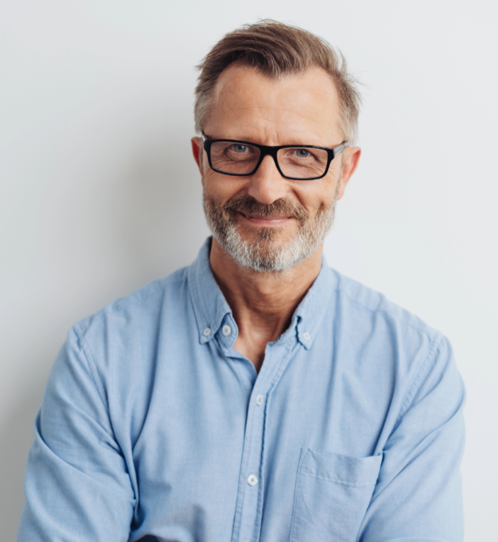A middle-aged man with gray hair and beard, wearing glasses and a light blue button-down shirt, smiling at the camera against a plain white background.