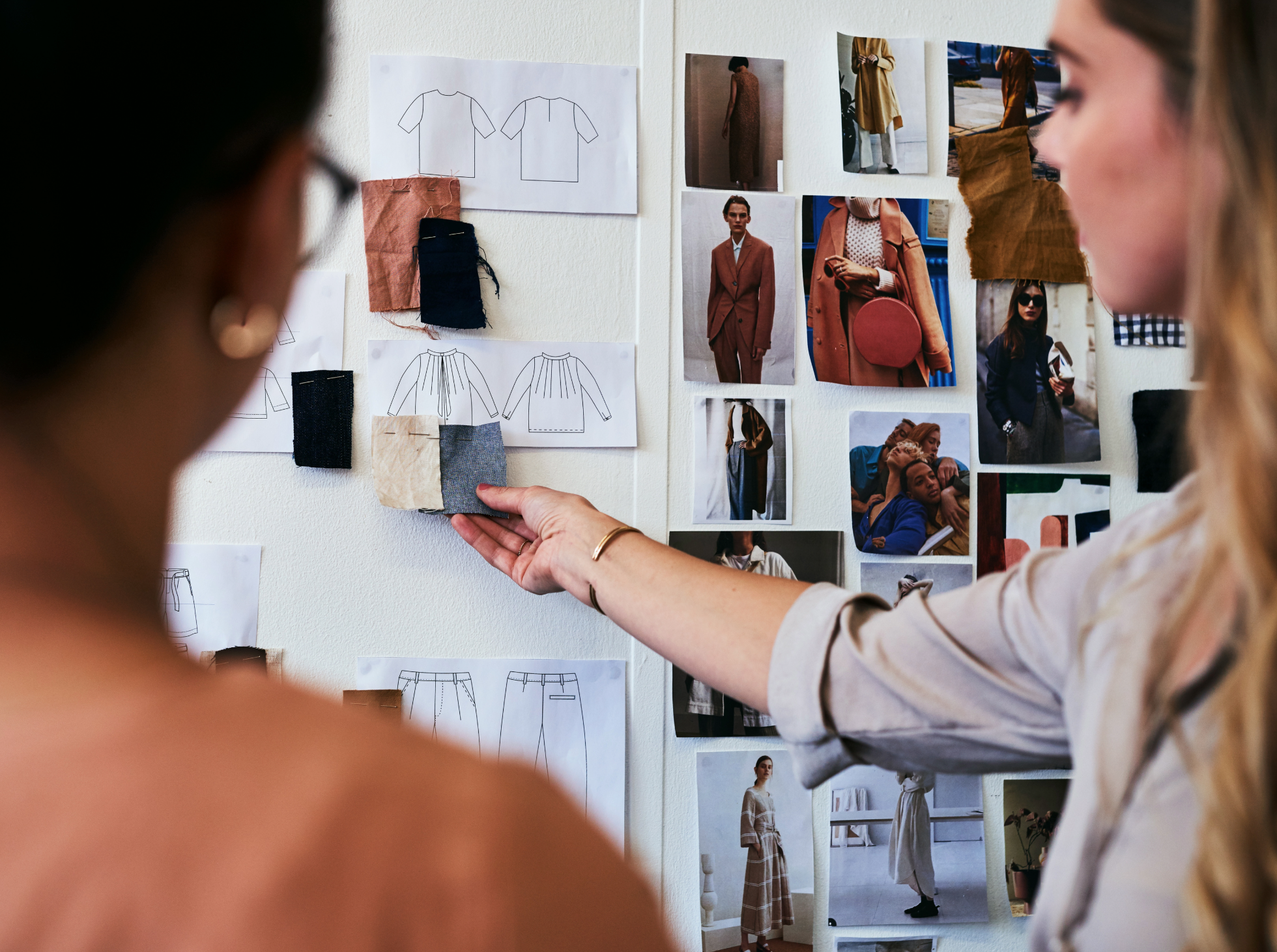 Two women looking at a wall with fashion sketches, fabric swatches, and photographs.
