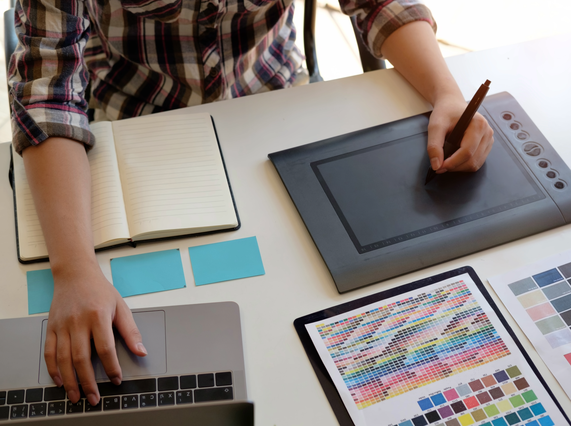 Person using a stylus on a graphics tablet at a workspace with a color palette guide and color swatches.
