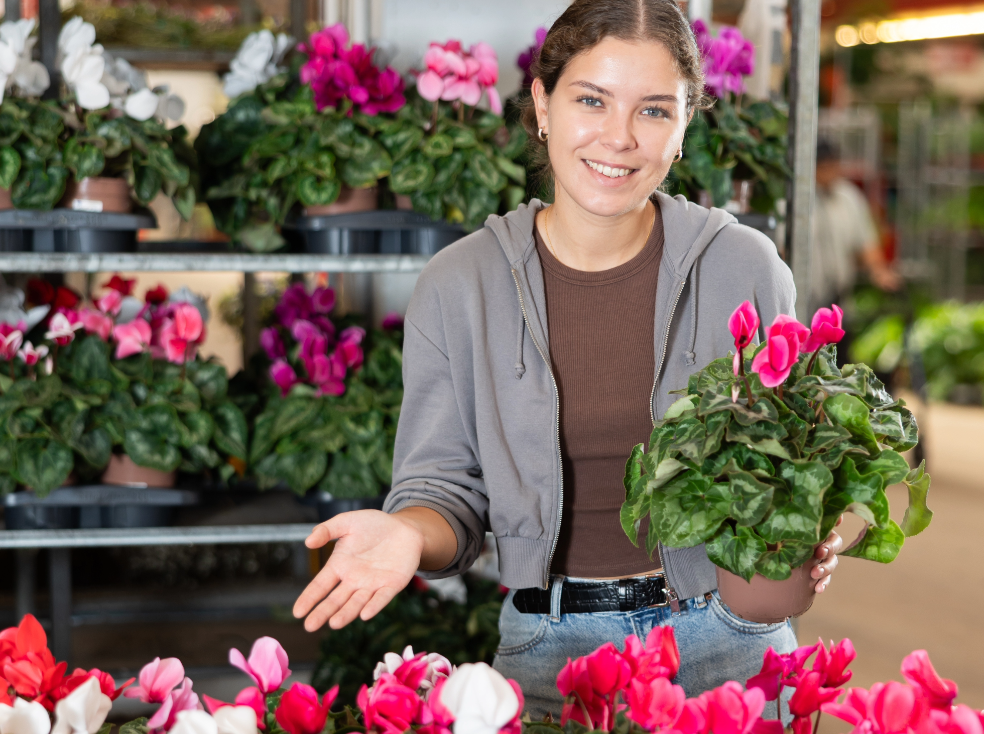A young woman smiling while holding a potted pink flowering plant in a greenhouse.
