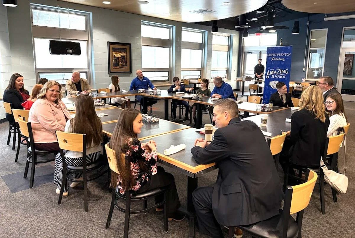 People sitting around tables in a room with large windows, listening to a speaker or presentation. There is a blue banner with text behind the speaker.