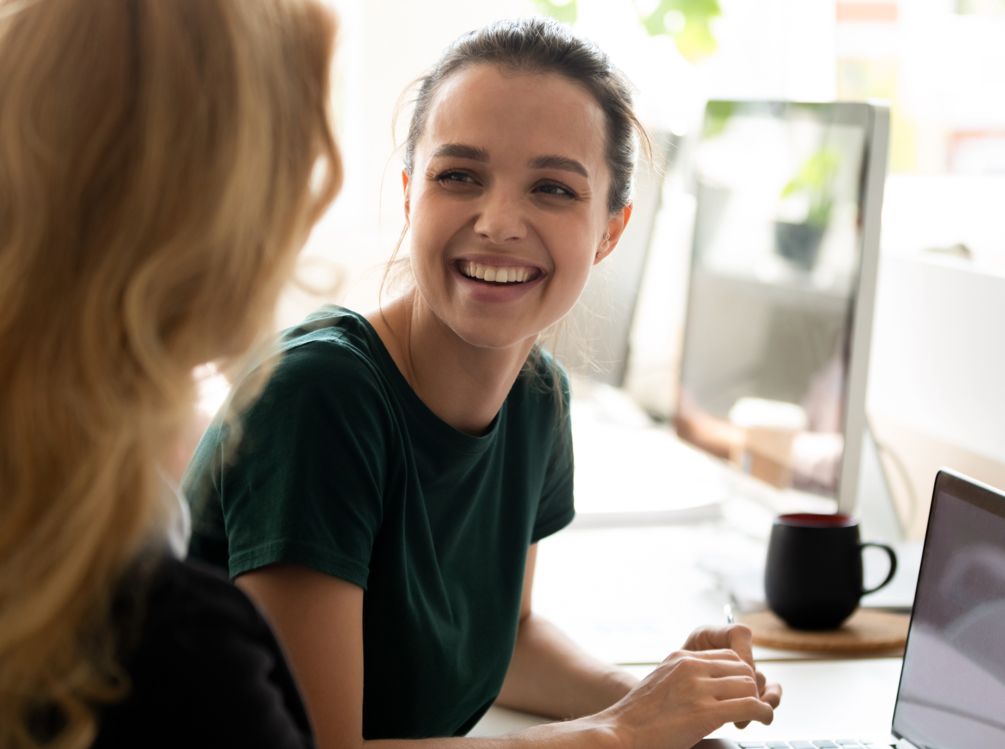 Two women sitting at a desk, laughing and talking, with a computer and a coffee mug in a bright, modern office.