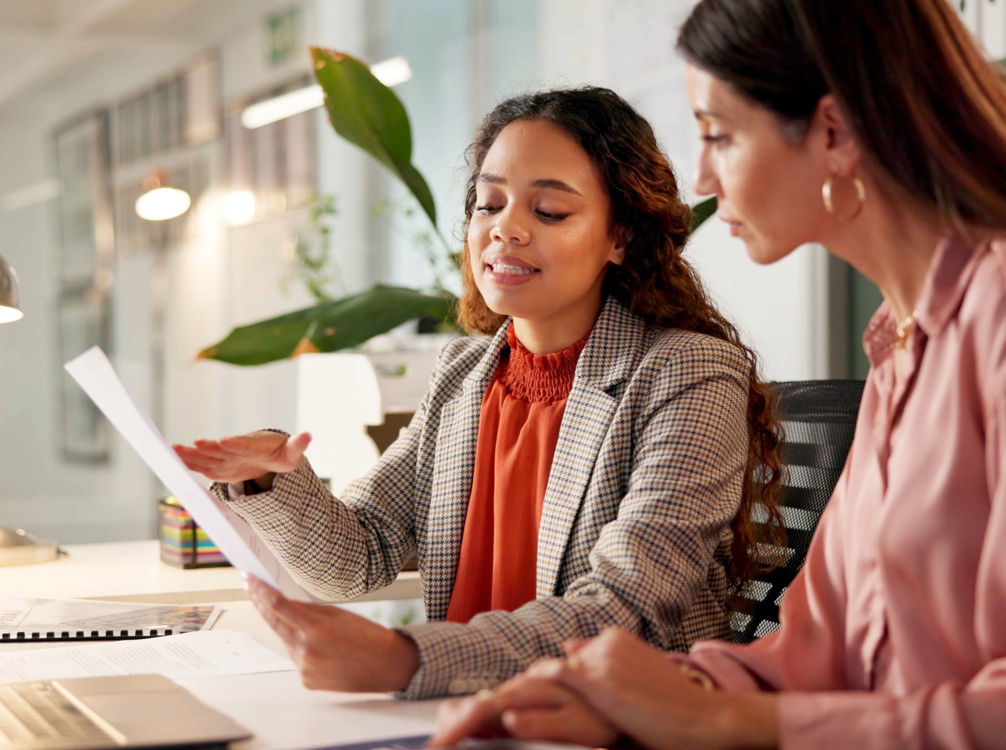 Two women sitting at a desk having a discussion, with documents and a laptop in front of them.