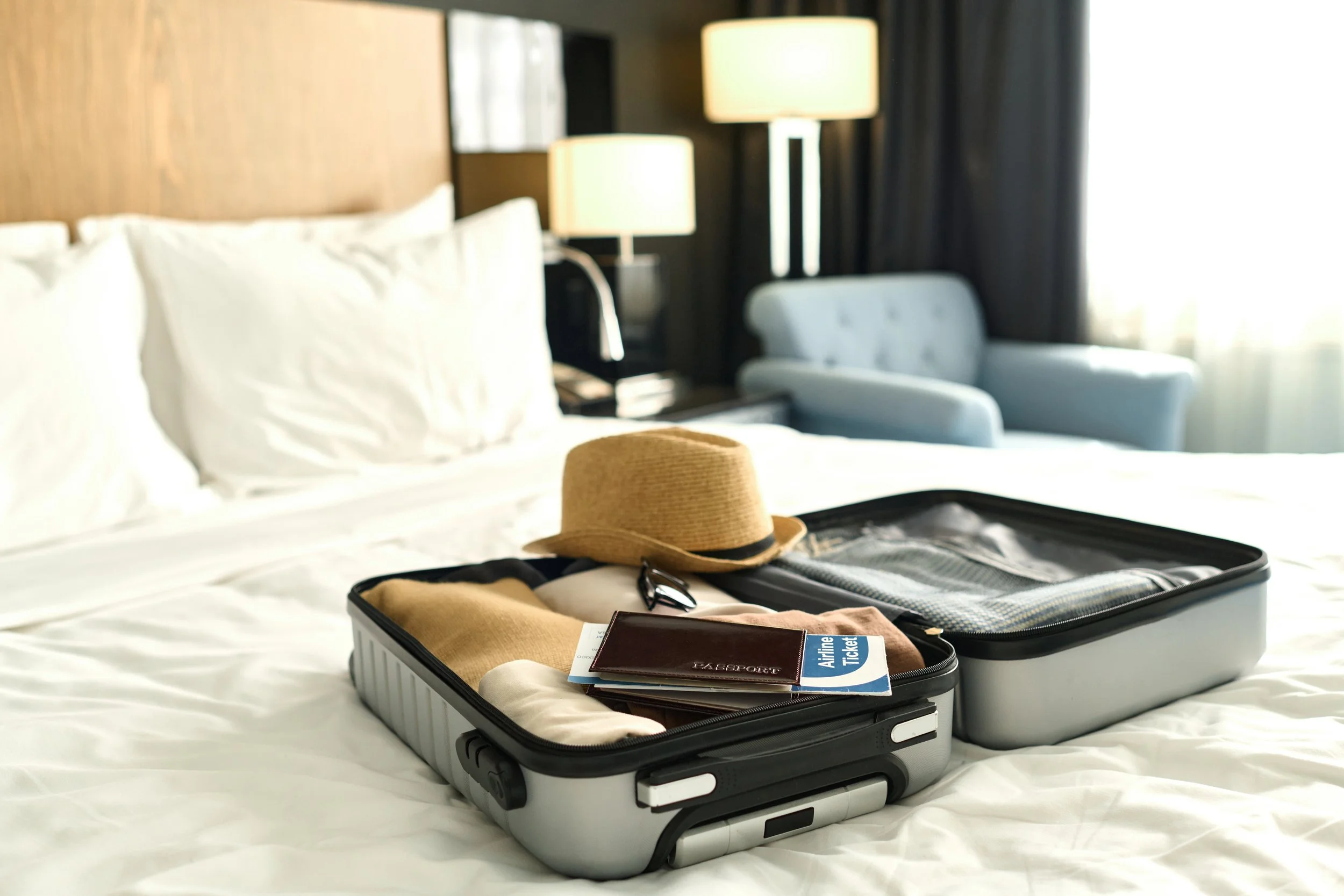 Open suitcase on a hotel bed containing a straw hat, a pair of sunglasses, a passport, an airline ticket, and clothing.