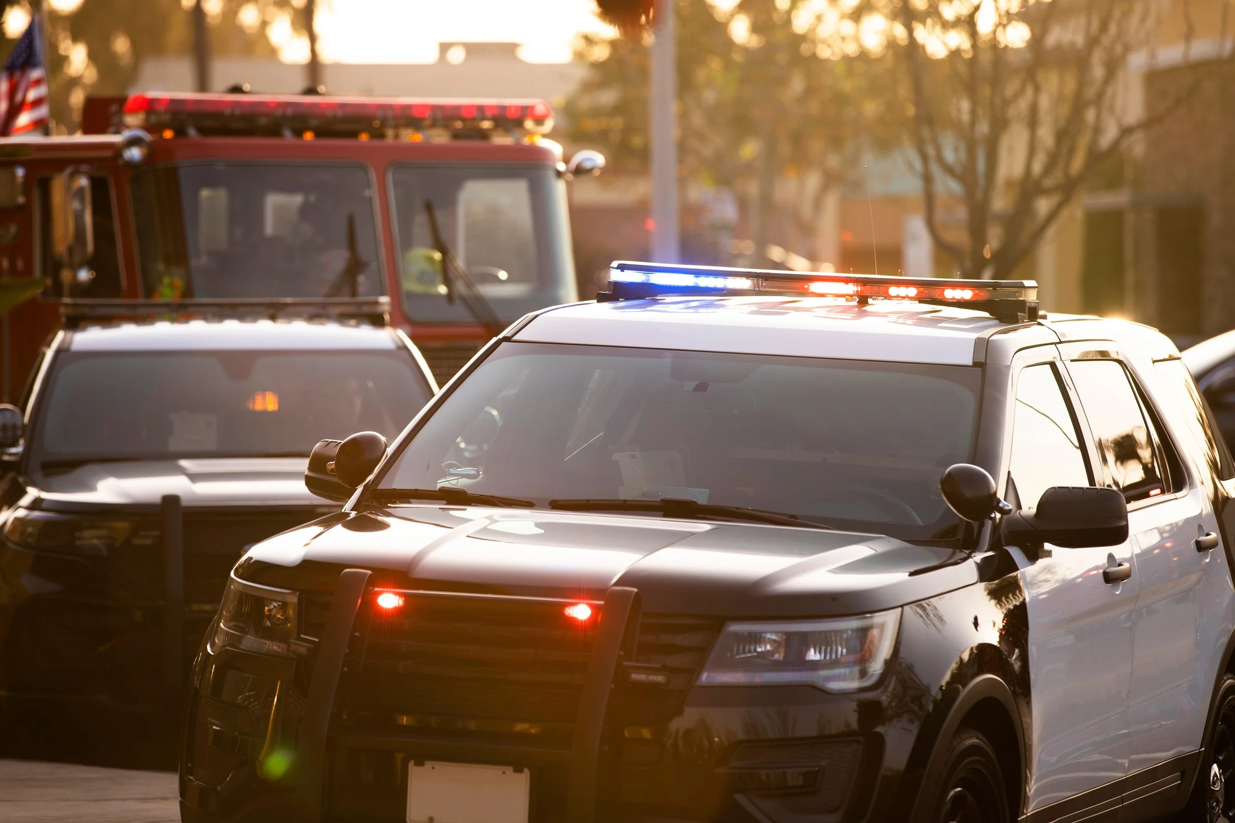 Multiple police cars with flashing red and blue lights parked on the street at sunset, with a fire truck visible in the background.