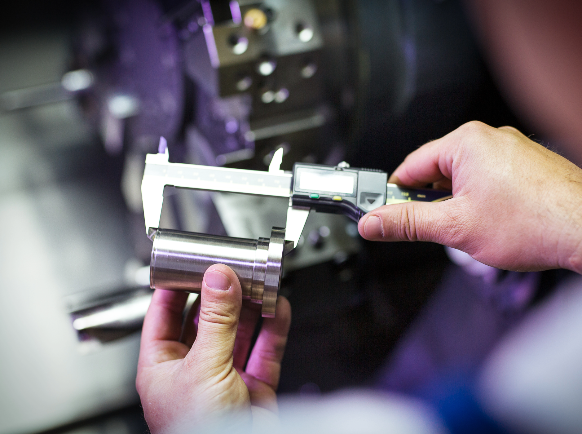 A person holding a digital caliper measuring a shiny metal cylindrical object in a industrial setting.