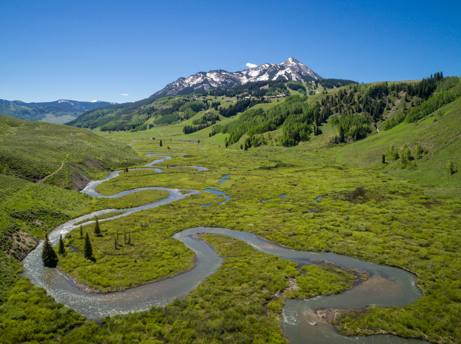 A winding river flows through green grassy hills with scattered trees, with snow-capped mountains and a clear blue sky in the background.