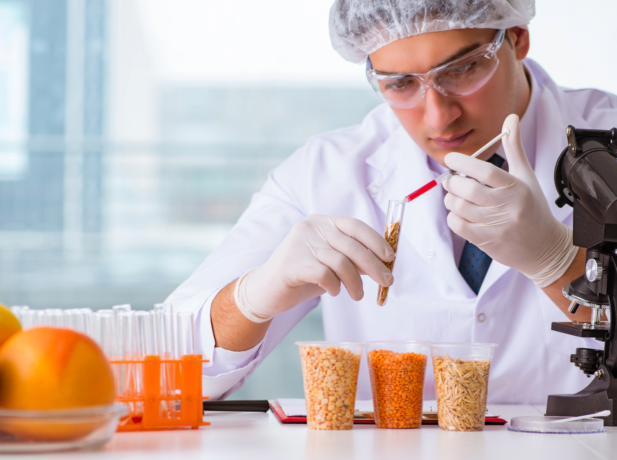 A scientist wearing safety goggles, gloves, and a hairnet, working in a food testing facility. The person is using a pipette to transfer a liquid into a test tube, with various containers of dried food samples and a microscope nearby.