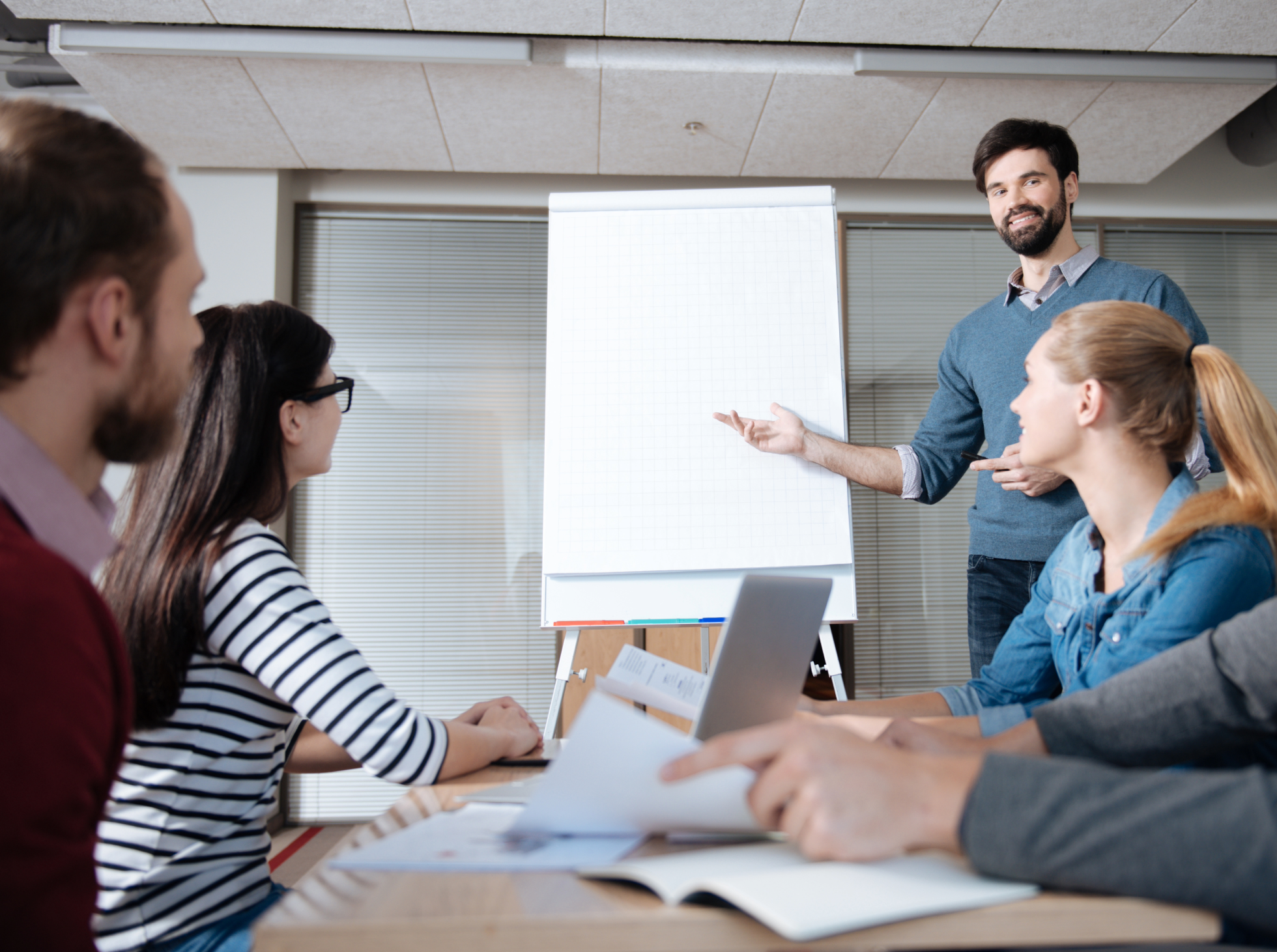 A man presenting to a group of four women seated at a table, in a classroom or conference room with a flip chart.