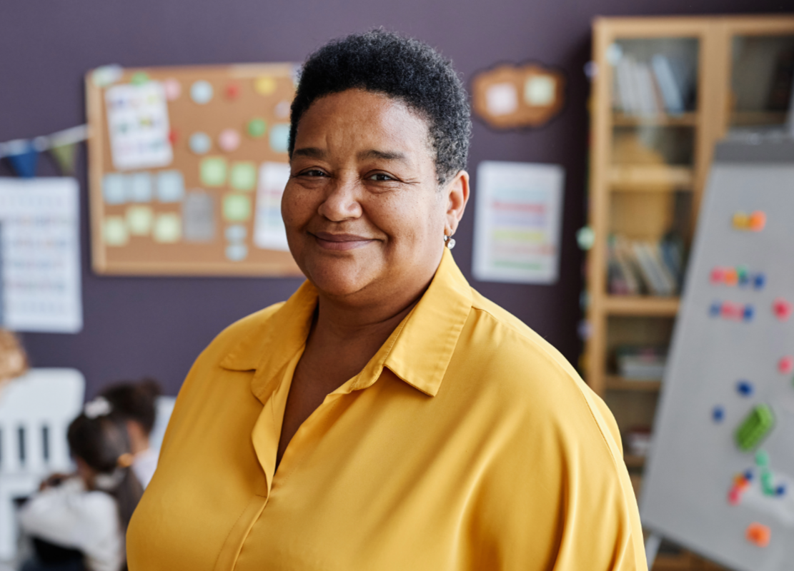 A smiling woman in a yellow shirt standing in a classroom with children in the background.