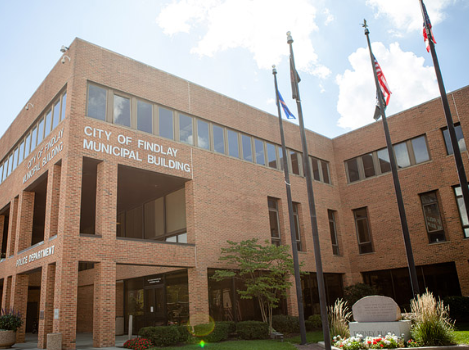 The City of Findlay Municipal Building, a multi-story brick government building, with several flagpoles displaying flags, including the American flag, in front.
