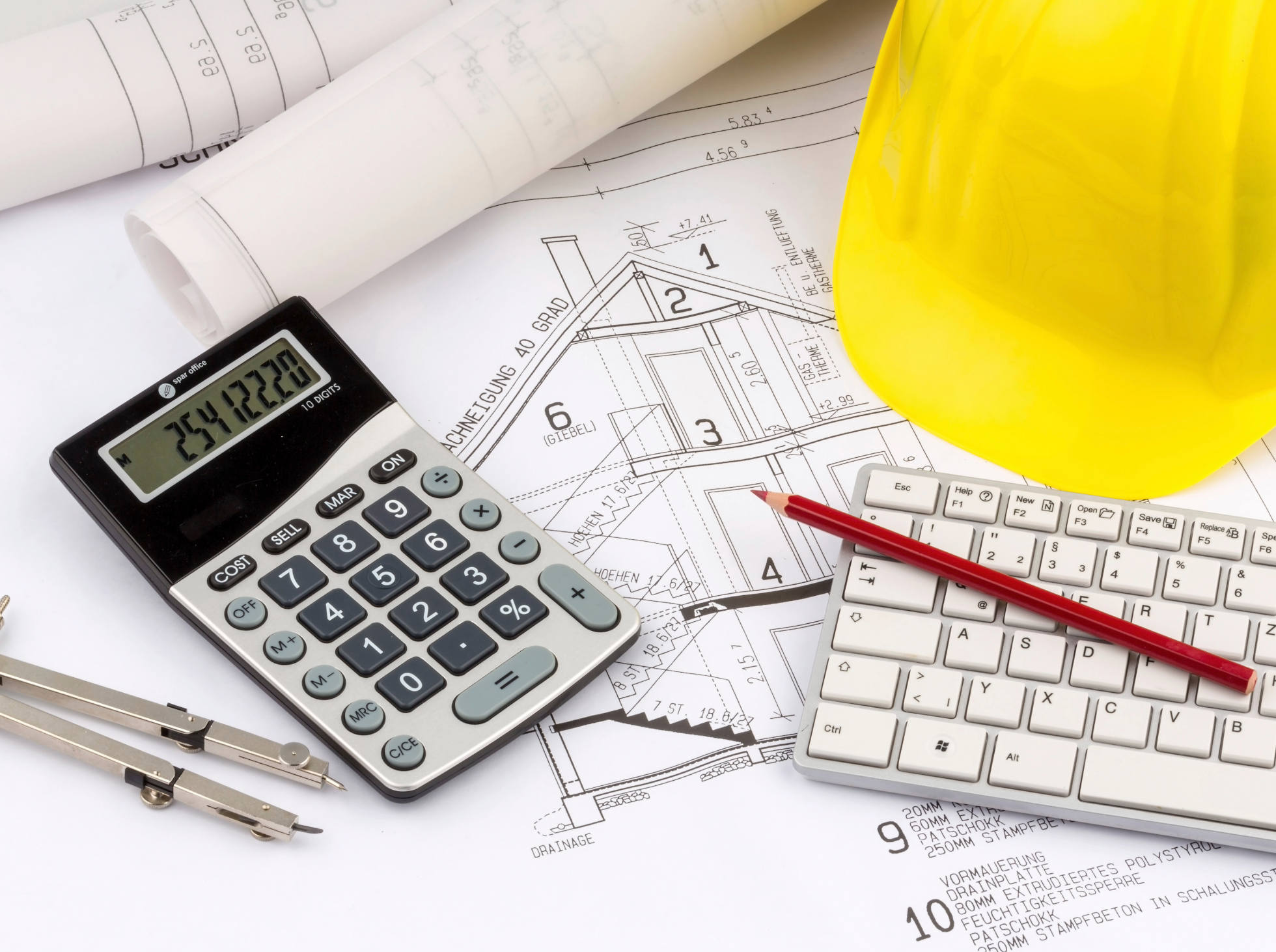 A yellow safety helmet, a calculator, a red pen, a white keyboard, and architectural blueprints with measurements and sketches in an engineering or construction workspace.