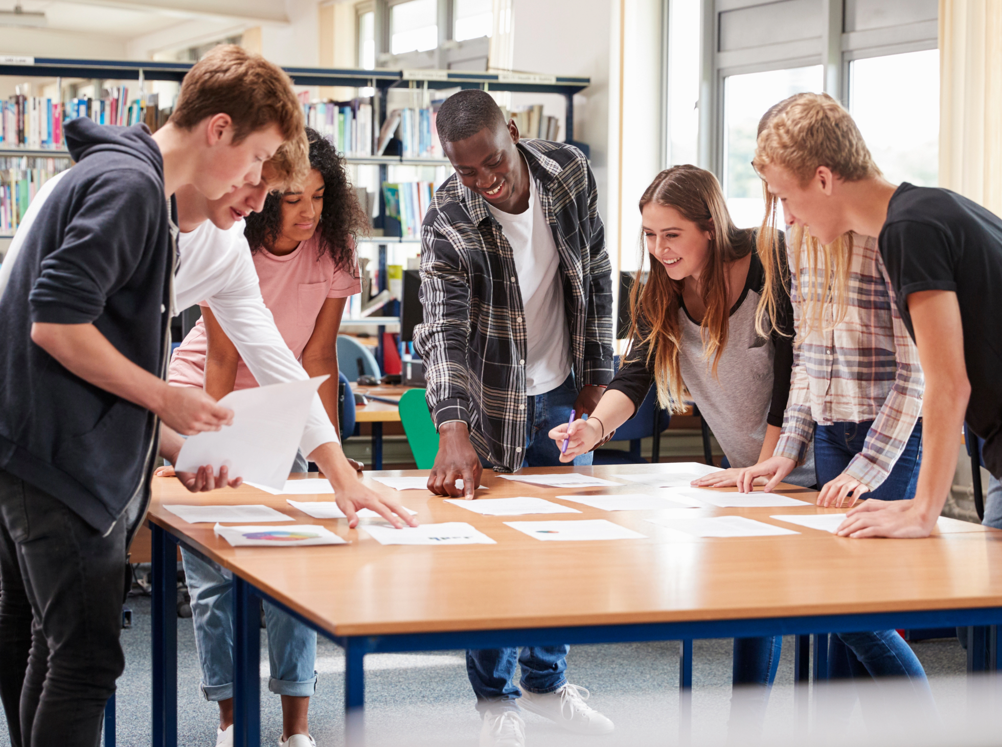 A group of six diverse students leaning over a table in a library, working together on papers and notes, smiling and engaged.