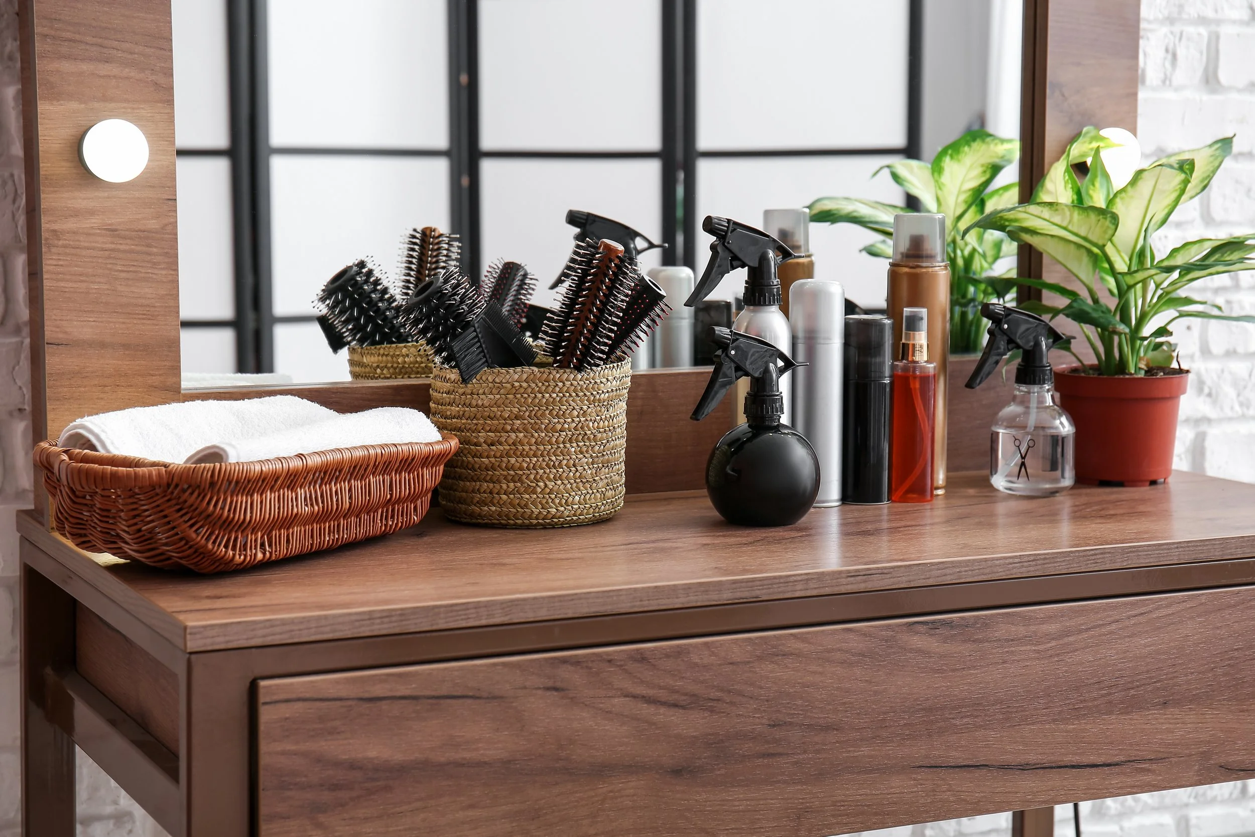 A hair salon table with a mirror, holding hairbrushes, spray bottles, and potted plants, with a white towel in a woven basket in the foreground.