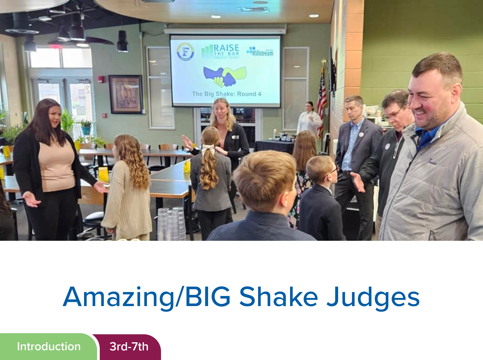 A group of people, including children and adults, are gathered in a conference room for an event titled "The Big Shake: Round 4". The room has a large screen displaying the event logo and title, with a handshake graphic. Some people are smiling and conversing. The setting appears professional and engaging, with participants participating in a discussion or presentation.