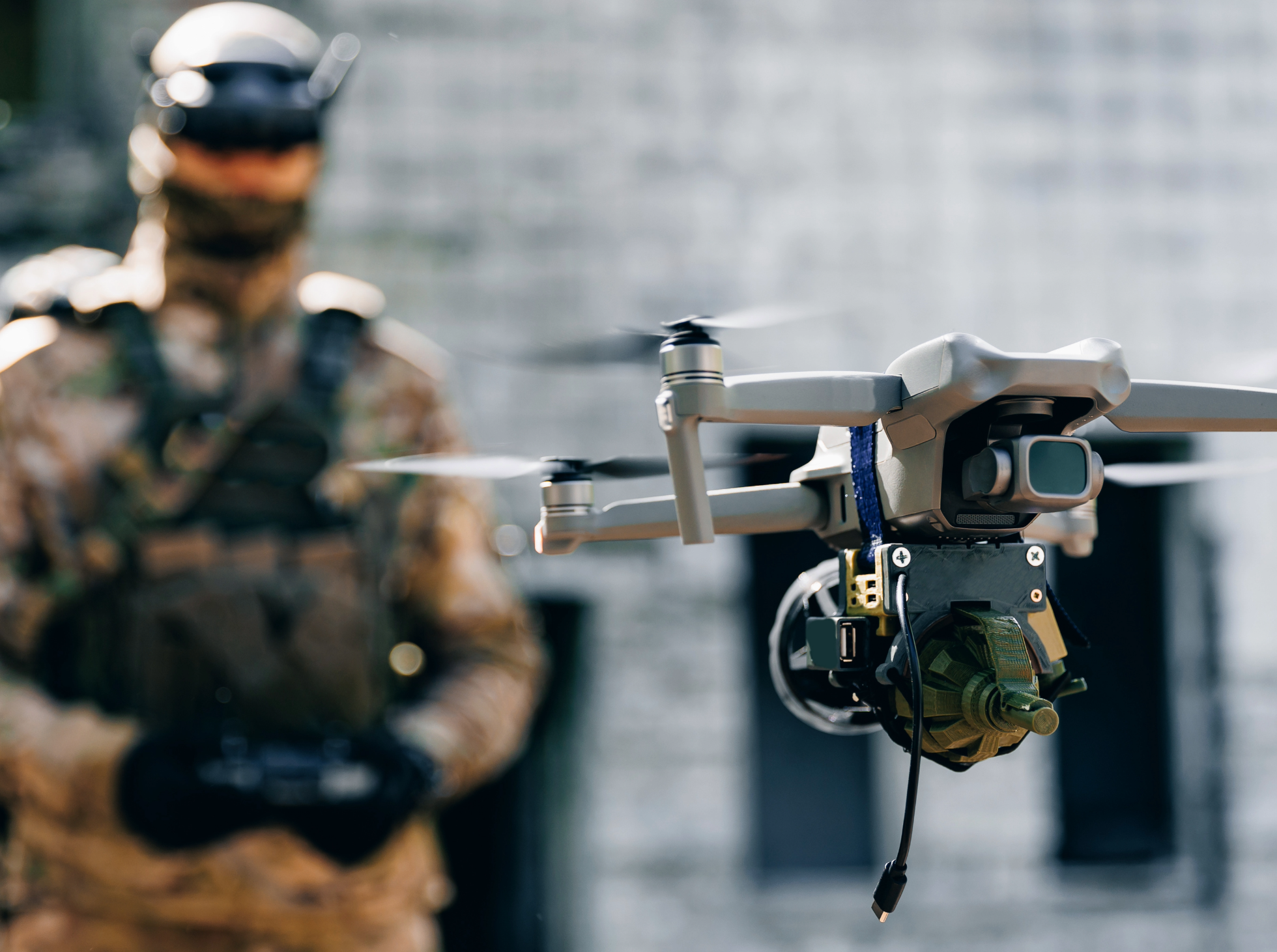 Close-up of a drone equipped with a camera and a robotic arm, with military member in the background.
