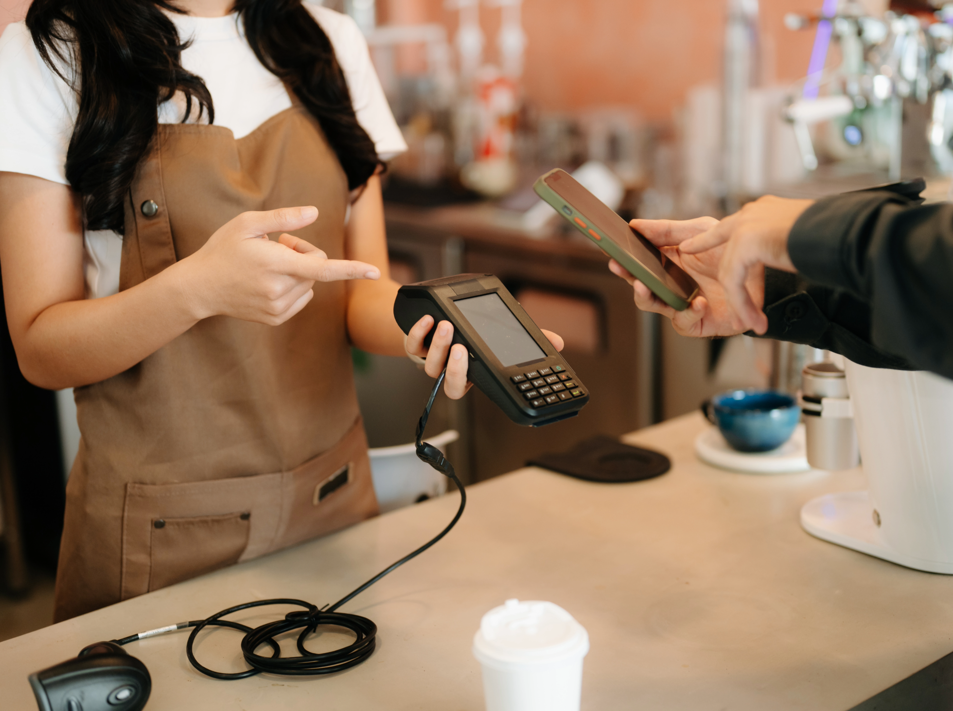 A woman wearing a brown apron is in a cafe or coffee shop, interacting with a person holding a smartphone and a card terminal making a payment. There are coffee cups and other items on the counter.