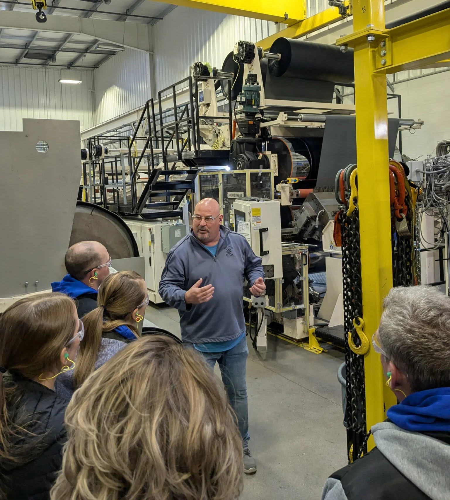 A tour guide is explaining machinery to a group of people in an industrial warehouse, with various large machines and equipment visible in the background.