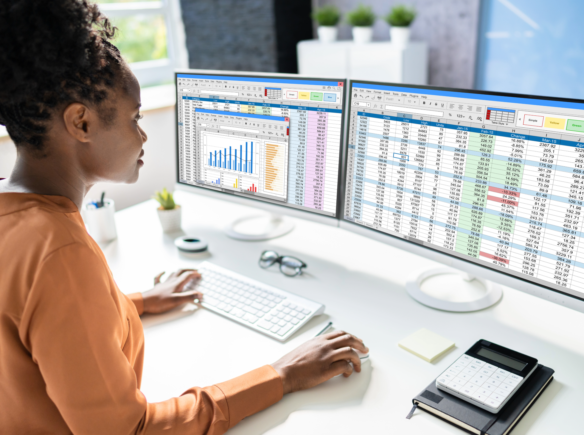 A woman sitting at a desk working on two computer monitors displaying spreadsheets with data and charts.