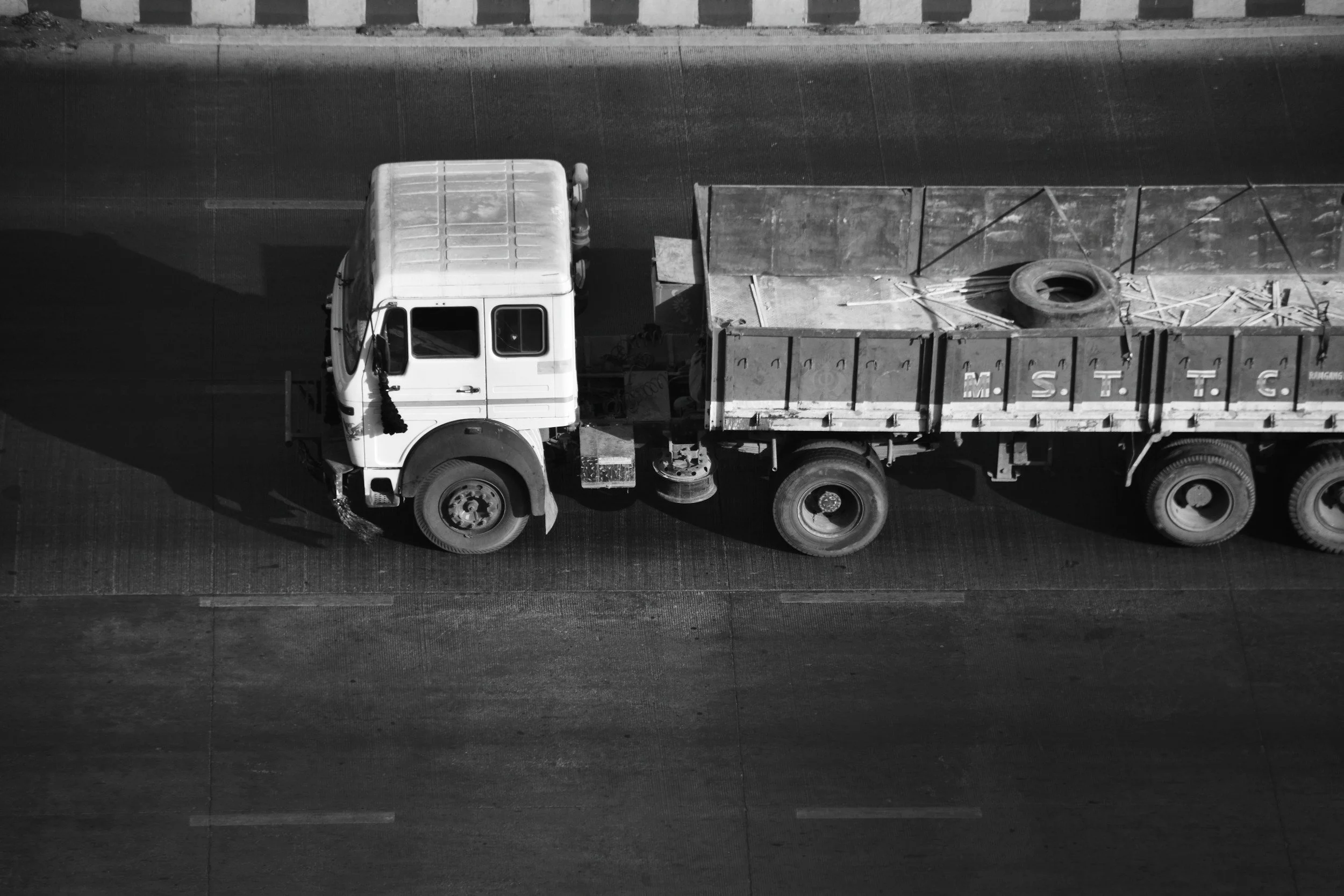 An aerial view of a large truck with a white cab and an open flatbed trailer on a highway. The flatbed is loaded with various construction materials, including a tire and wooden planks. The truck has a shadow cast on the road.