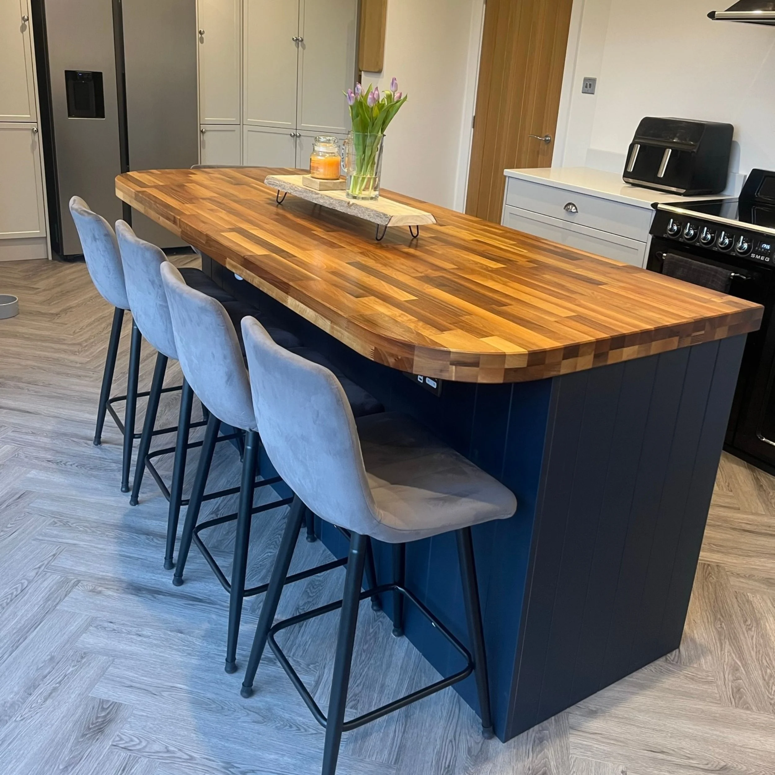 Kitchen island with a wooden top, four gray barstools, a flower vase with tulips, and a candle, surrounded by white cabinets and appliances.