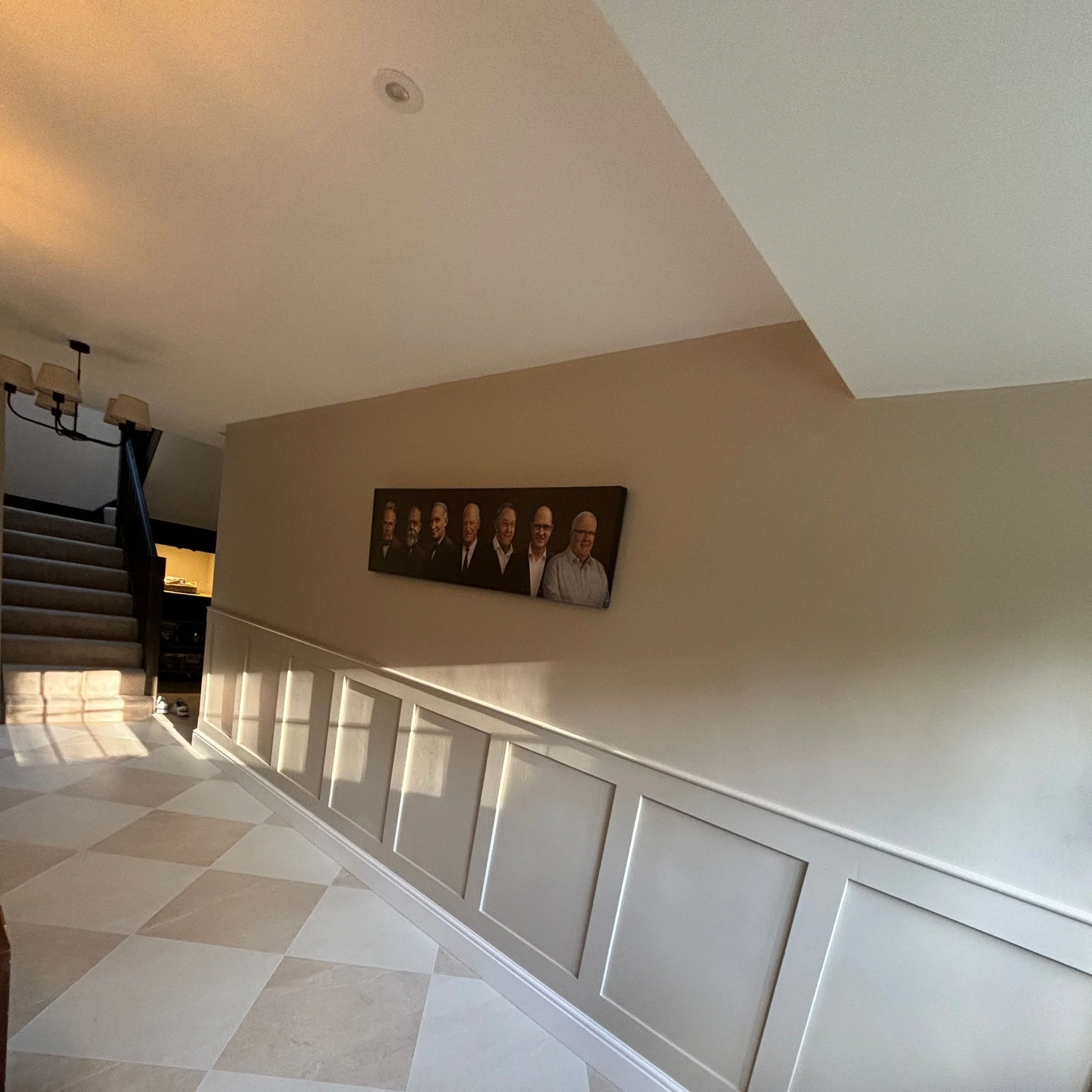 Interior of a home featuring staircase, wall decor with a framed group photo of men in suits, beige wall with white paneling, and tiled floor.