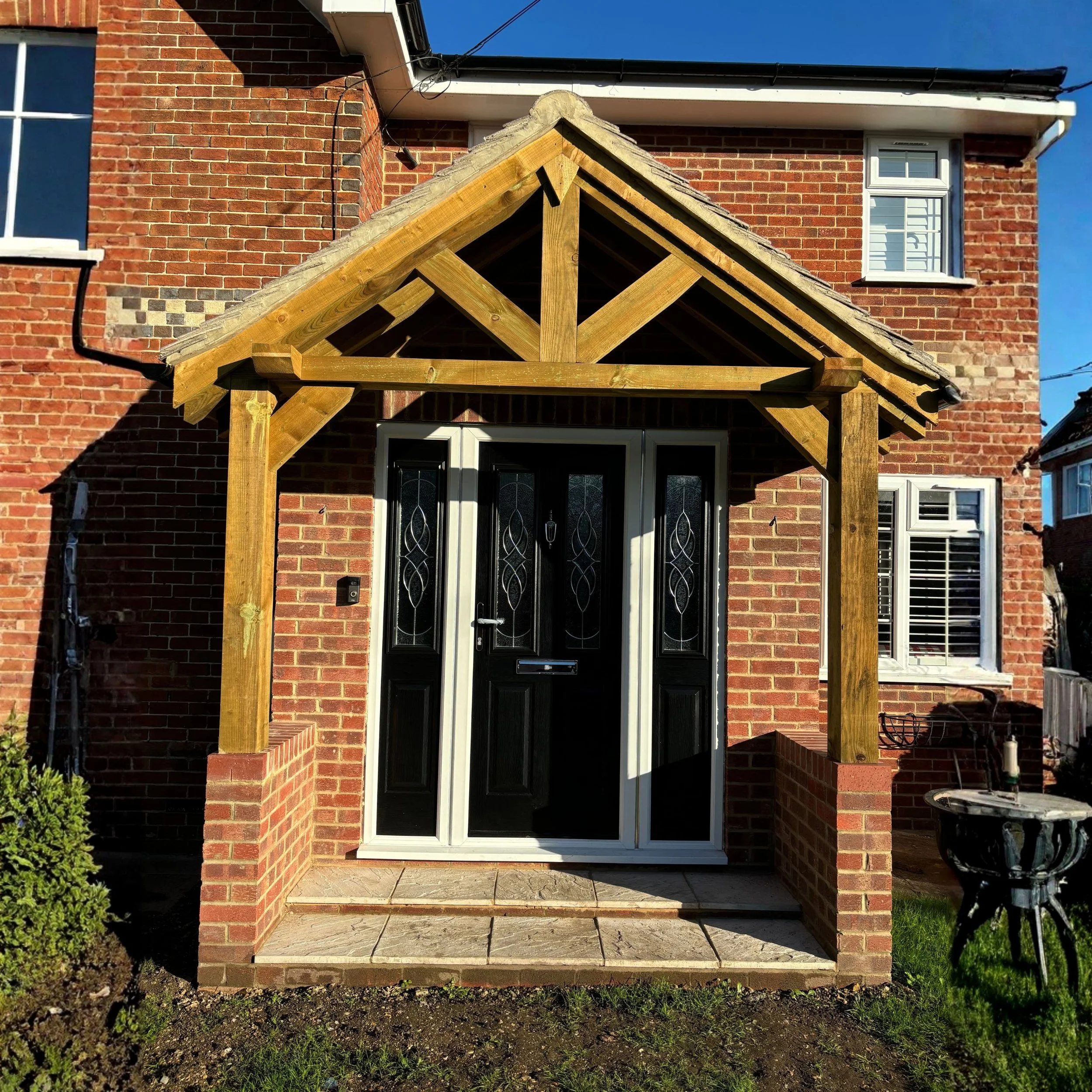 A new wooden porch frame is being built in front of a black front door with decorative glass panels on a brick house, with a small tiled platform in front and a garden on the side.