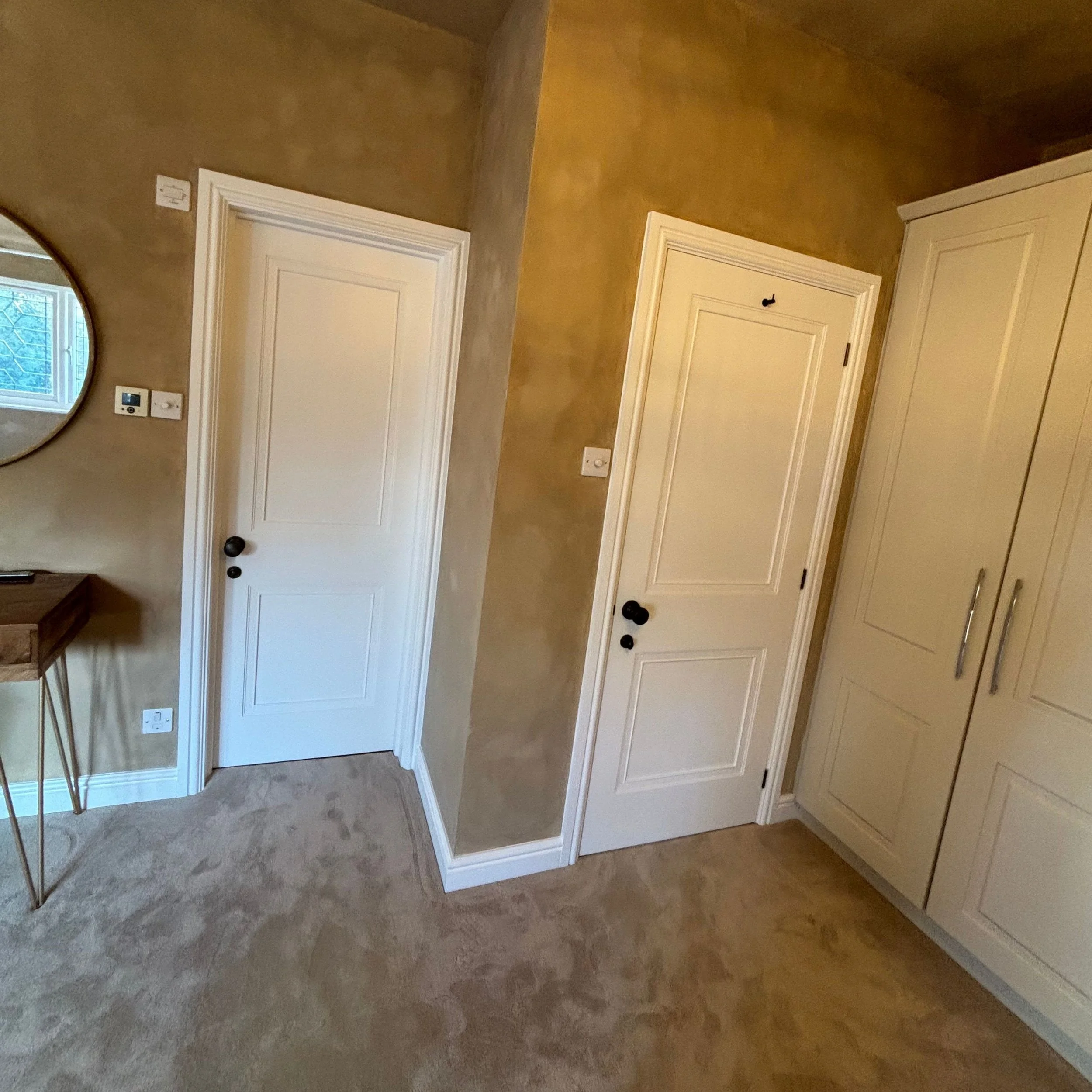 Two closed white doors with black knobs in a beige wall room, with a beige carpeted floor and wooden cabinet with silver handles on the right.