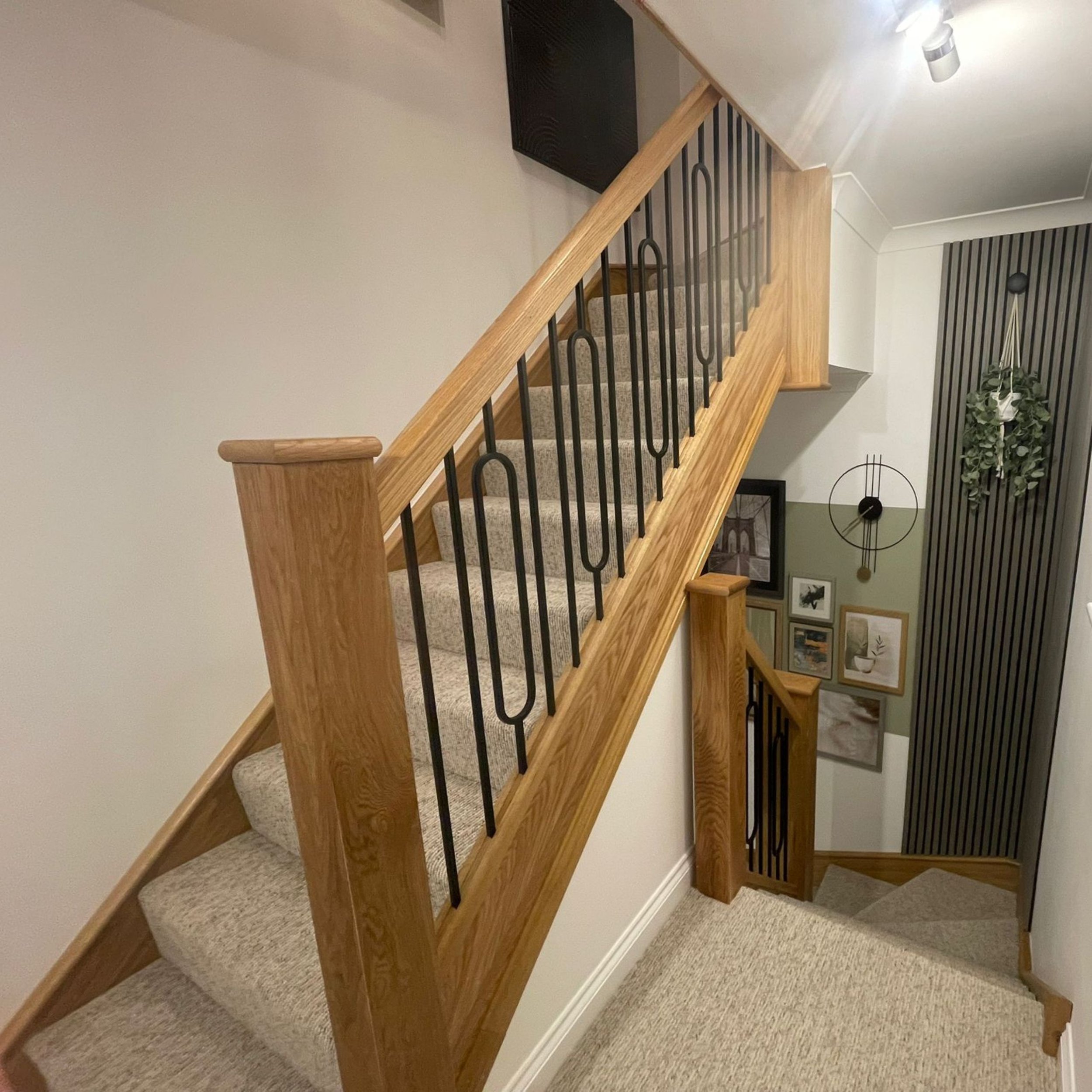 Wooden staircase with beige carpet, black metal railings, leading to the upper floor in a modern home interior with framed pictures and a clock on the wall.