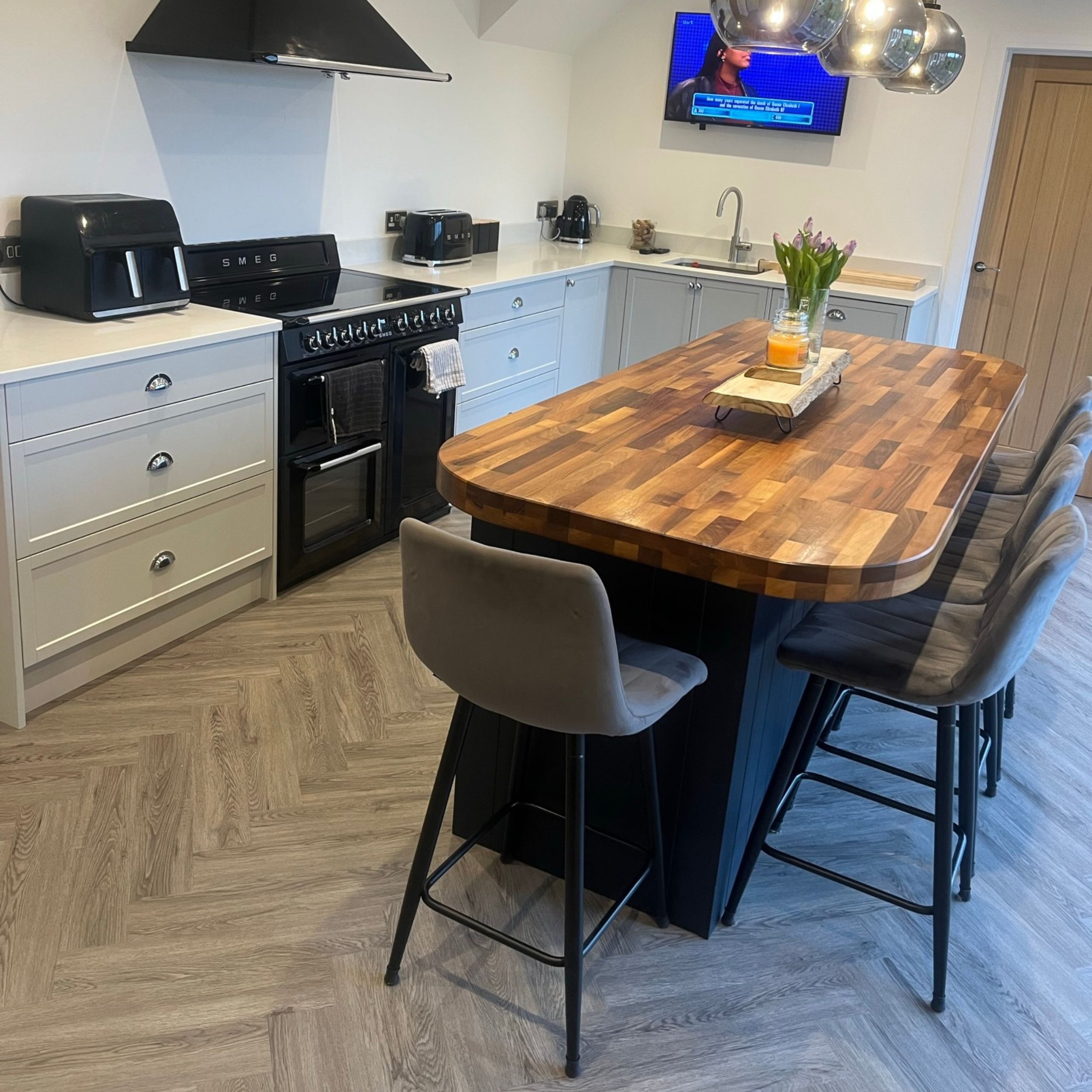 A modern kitchen with a large wooden island, gray cabinets, black appliances, and a wall-mounted TV. The island has a vase with tulips and a candle.
