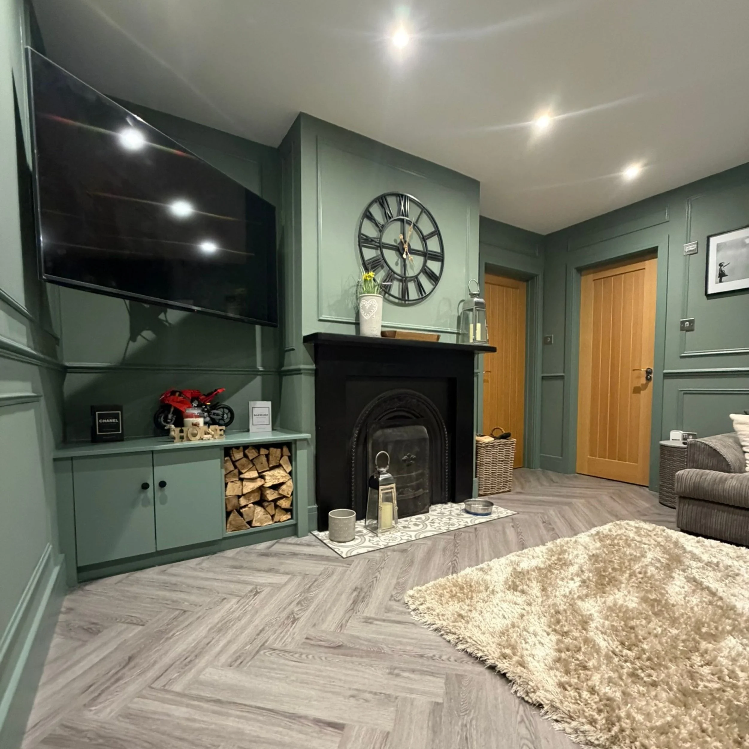 Living room with dark green walls, wooden doors, and a fireplace with black mantel. There is a large wall clock above the fireplace, a flat-screen TV on the left, and a light-colored fluffy rug on the floor. Decor items include candles, lanterns, and