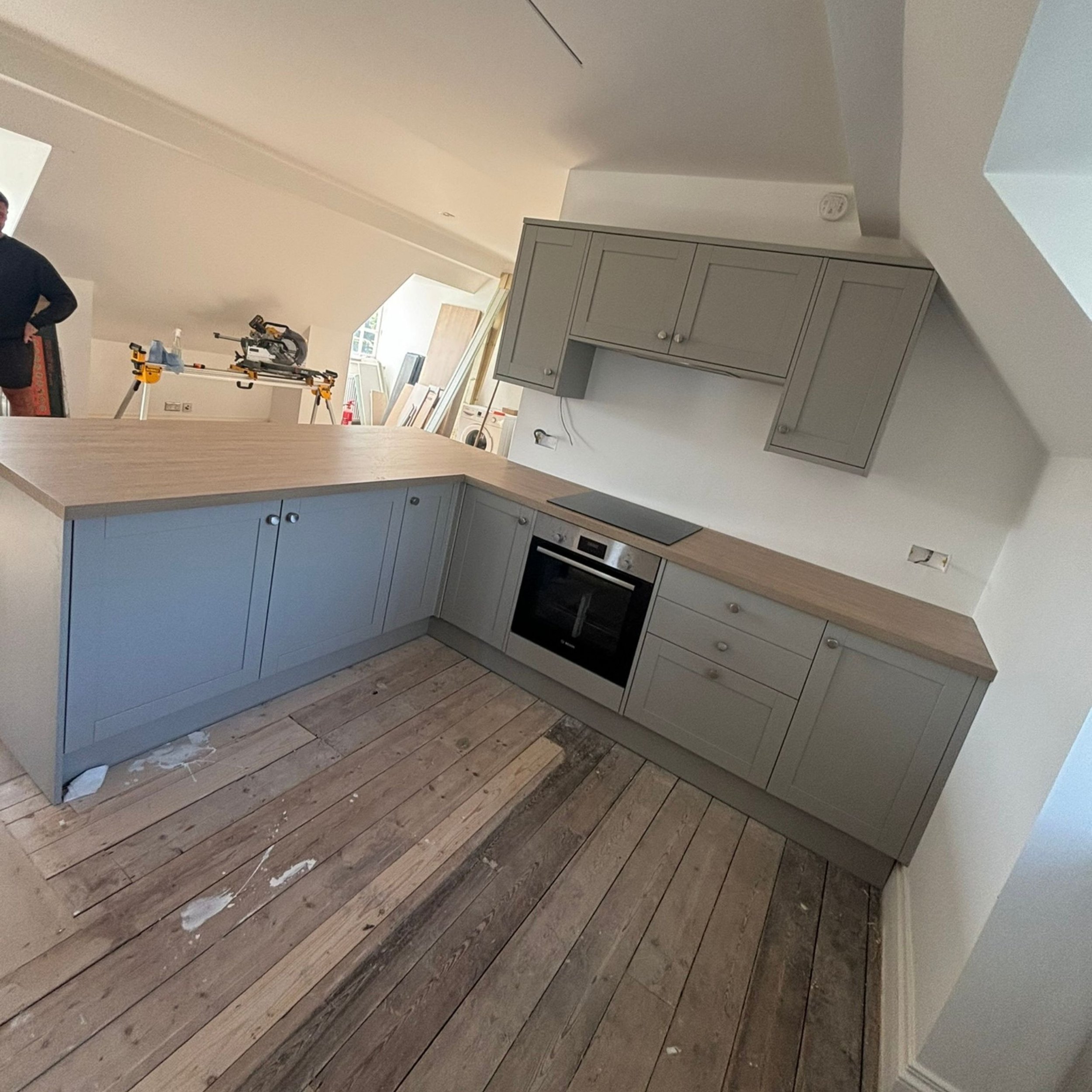 Unfinished kitchen with gray cabinets, a built-in oven, and a wooden countertop. There is construction equipment and a person in the background.