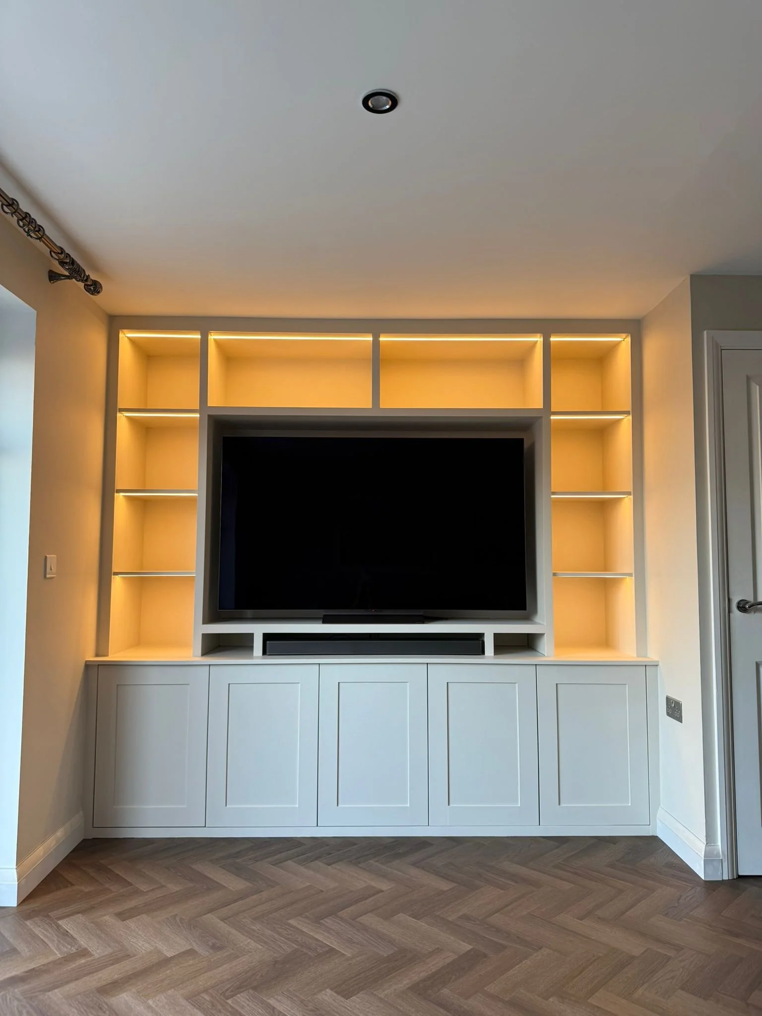 Living room with a black flat-screen TV on a white built-in entertainment center with open shelves and cabinets, and warm yellow lighting highlighting the shelves.