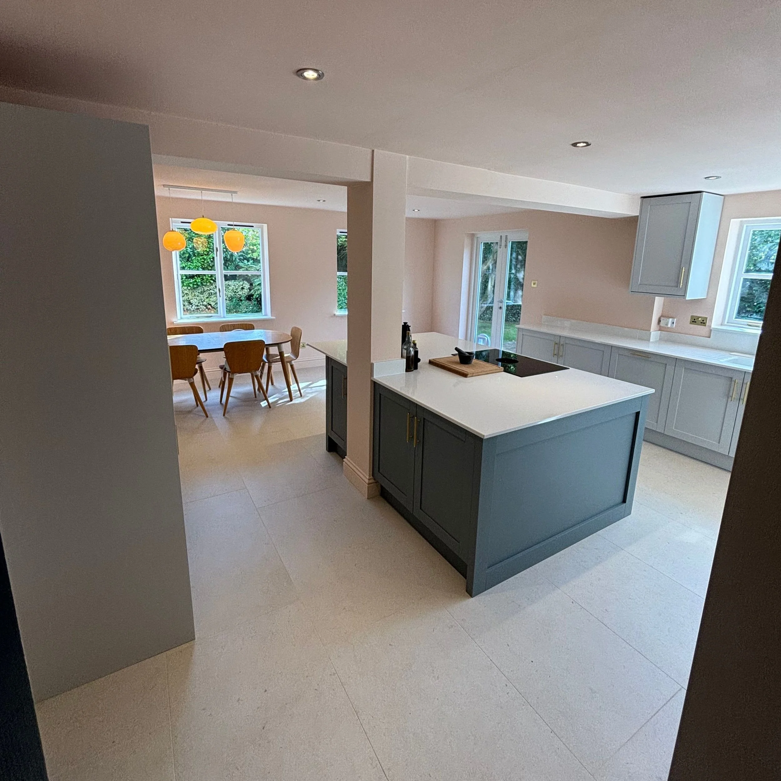 Modern kitchen with blue cabinets, white countertops, and a central island, adjacent to a dining area with a wooden table and chairs, and large windows and glass doors bringing in natural light.