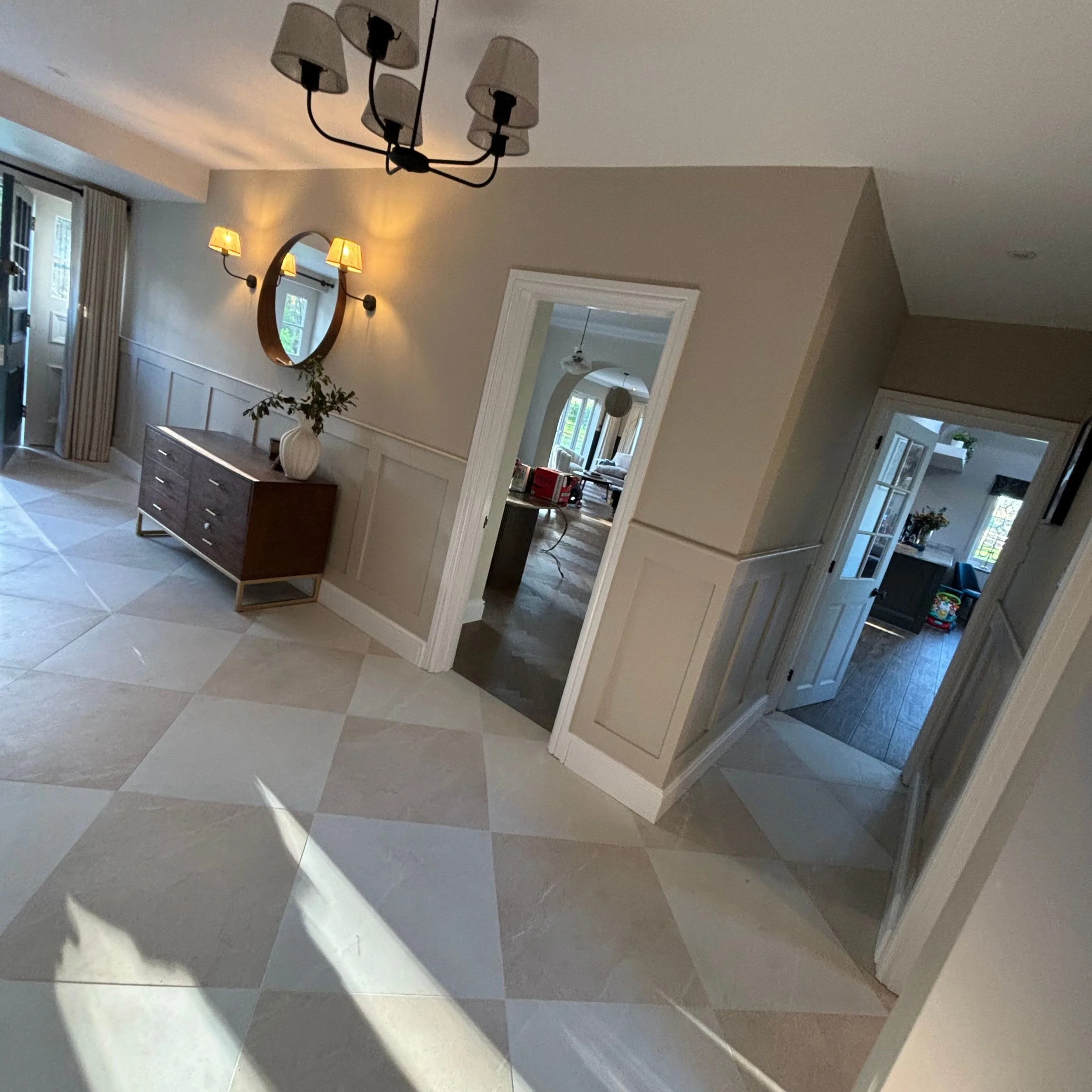 Interior view of a home with beige walls, a chandelier hanging from the ceiling, a mirror, a sideboard with a vase and plants, and multiple rooms visible through doorways.