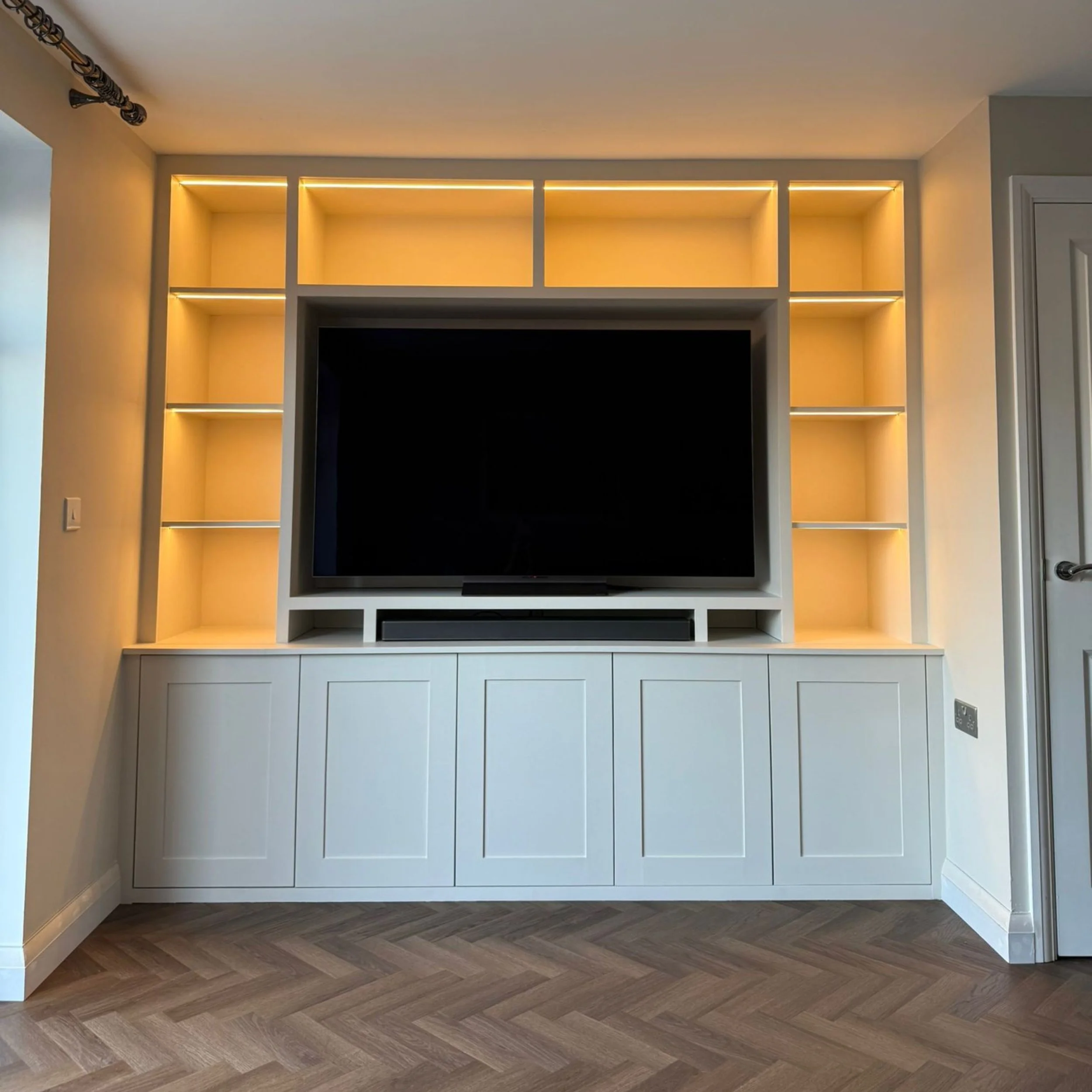 Modern white entertainment center with built-in shelving and a large flat-screen TV, illuminated with warm yellow LED lights, located in a living room with wooden chevron flooring.