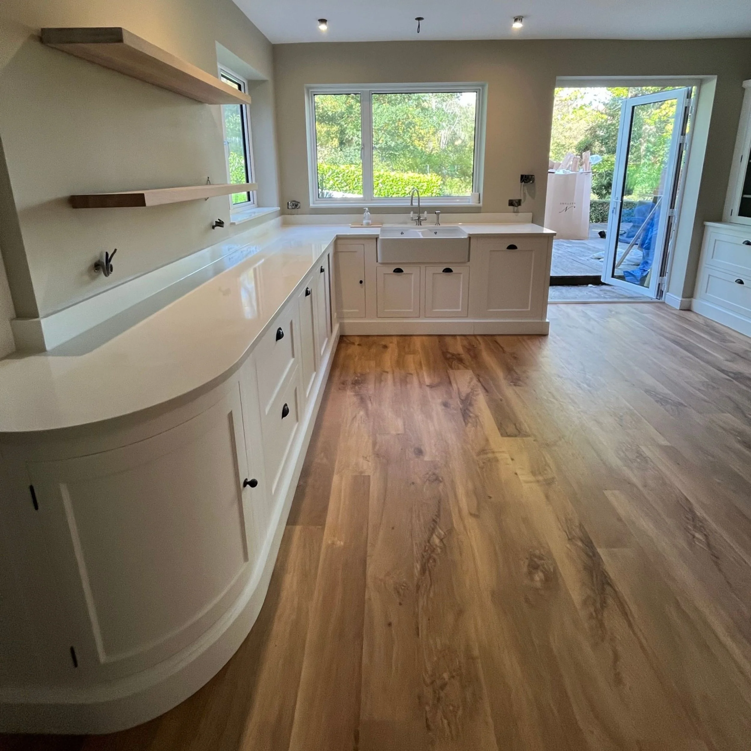 Empty kitchen with white cabinets, a large window, and a sliding glass door leading outside, with hardwood flooring and exterior view of green trees.