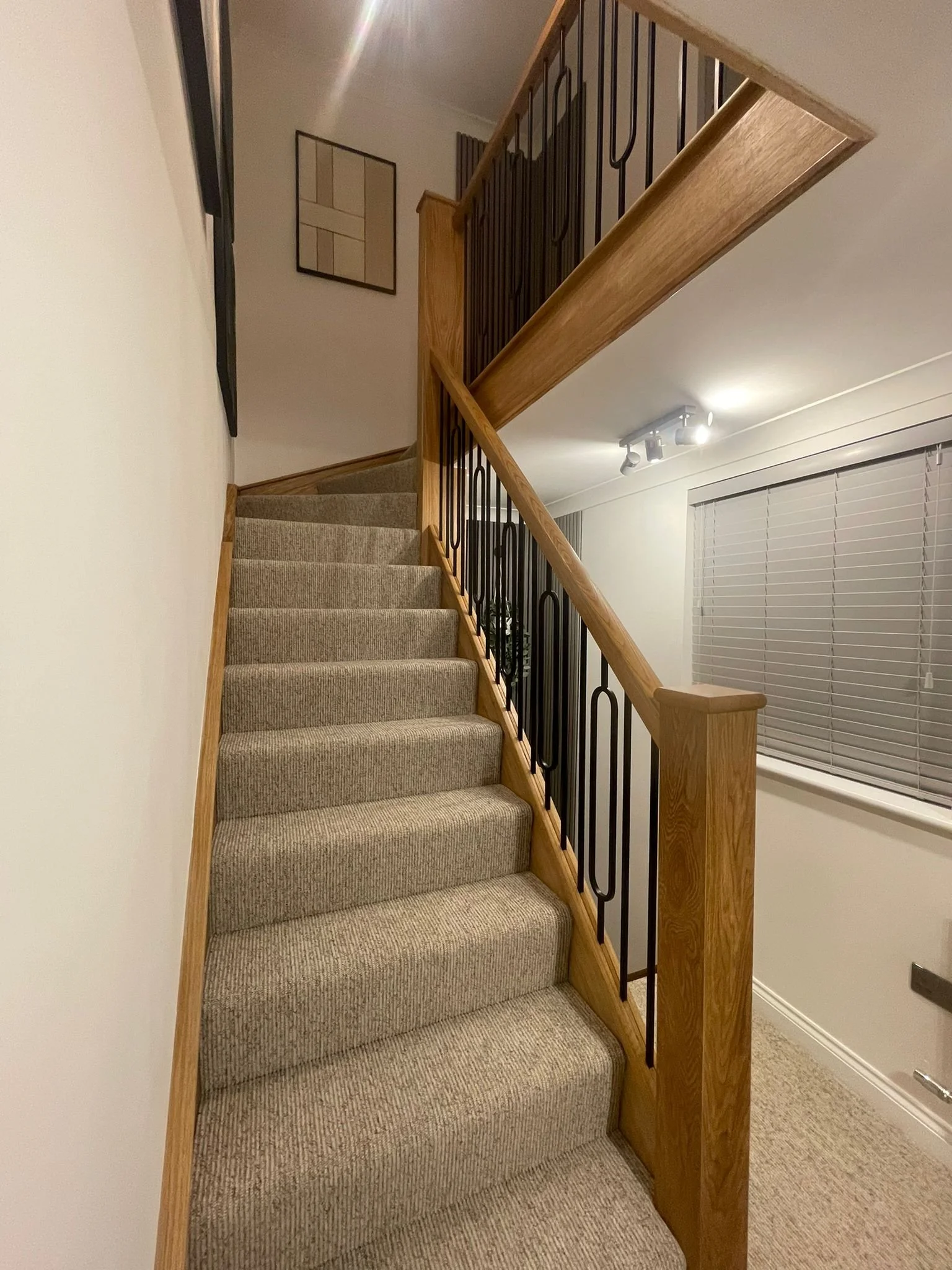 Interior view of a staircase with beige carpeting, wooden handrails, and black metal balusters, leading to the upper floor of a house or apartment, with wall art and a window with blinds on the right side.