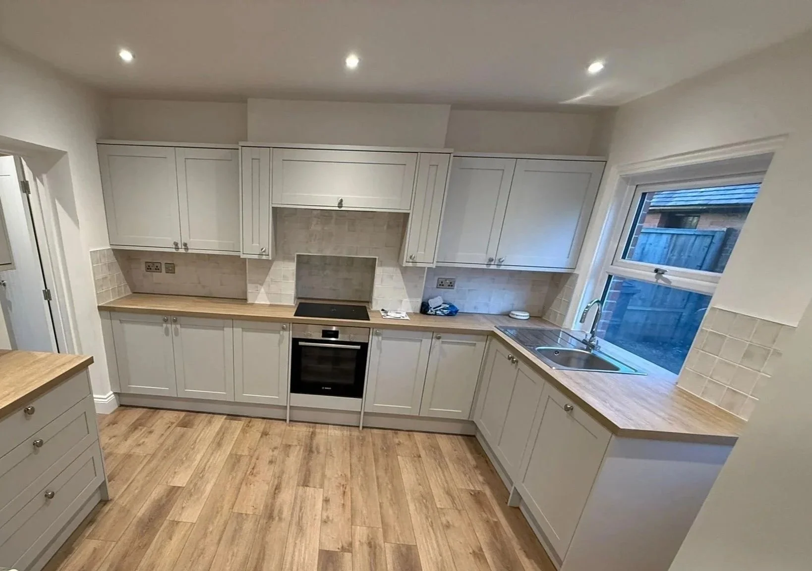 Kitchen with white cabinets, wooden countertops, a stainless steel oven, stovetop, and double sink under a window, with beige tiled backsplash and wooden floor.