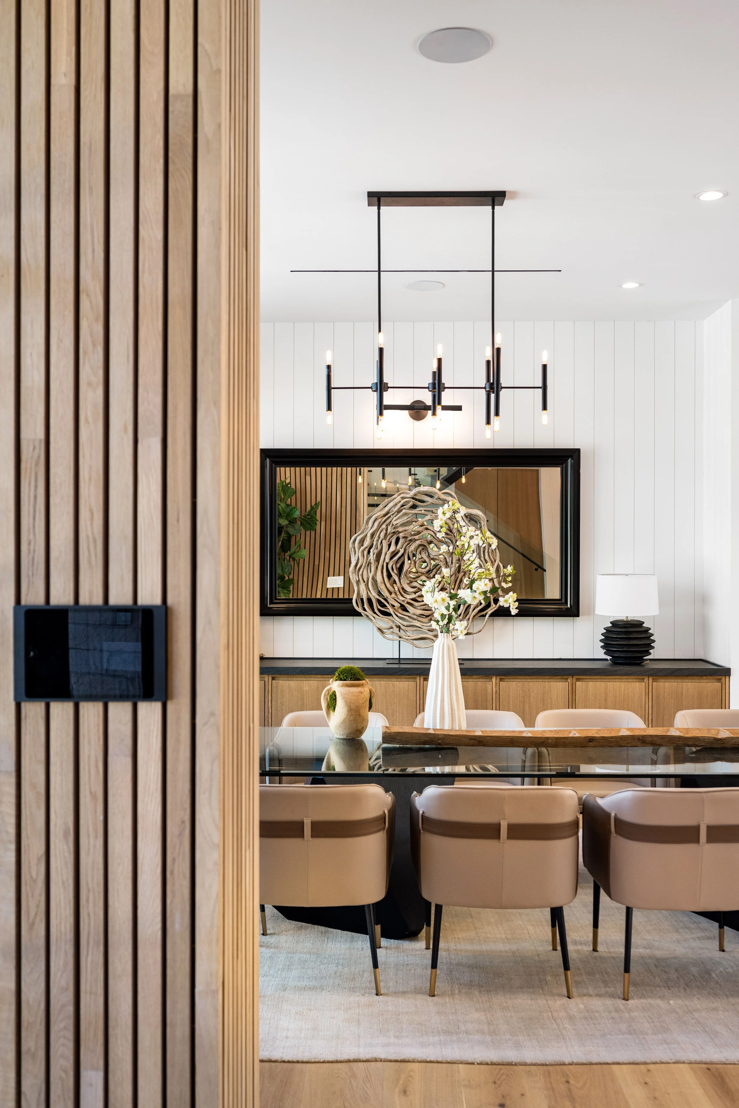 Interior of a modern dining room with a glass table, beige chairs, a large decorative floral arrangement, a mirror, and a wooden accent wall.