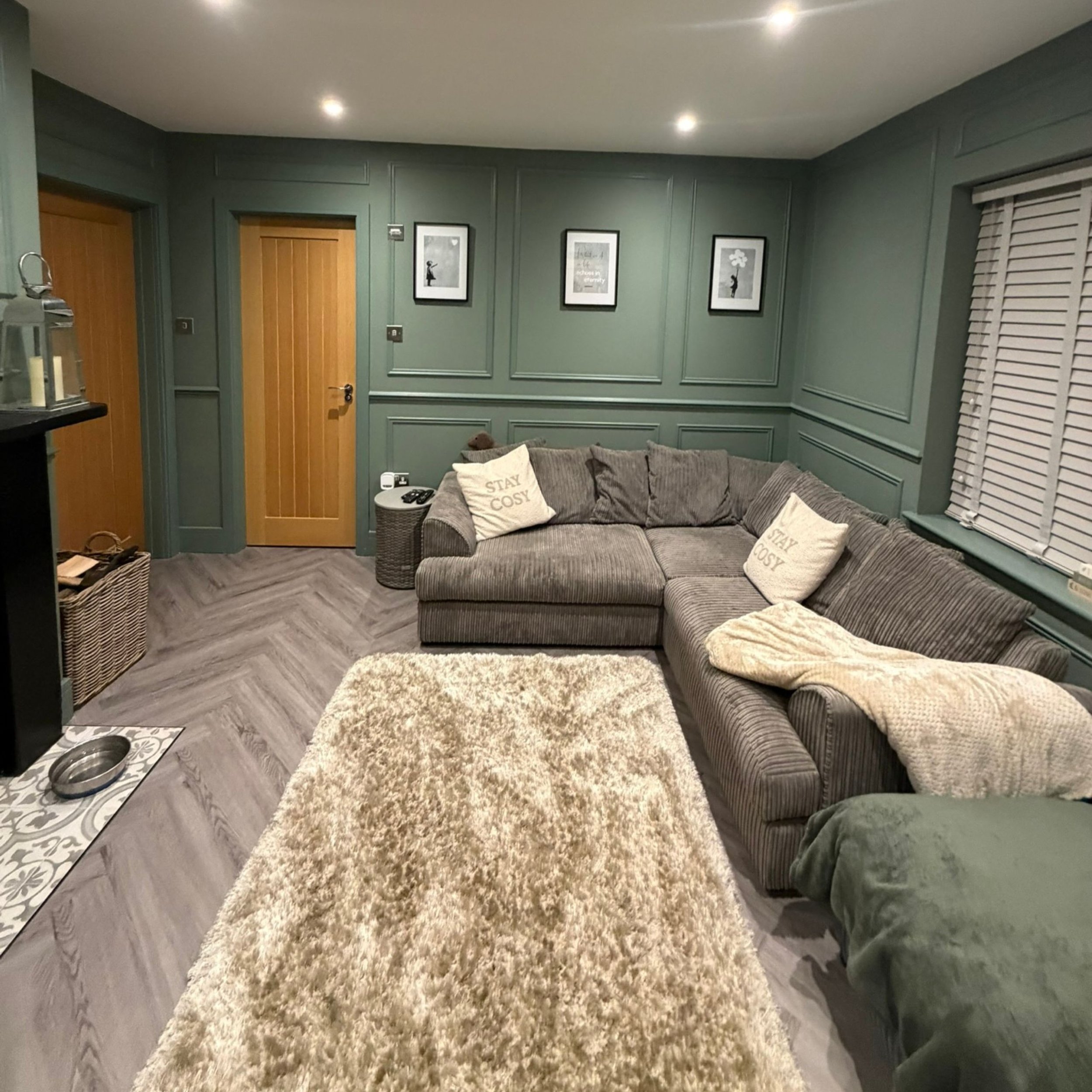 A cozy living room with green paneled walls, framed art, a large gray sectional sofa with pillows that say 'STAY COSY', a beige shaggy rug, a window with white blinds, and wooden doors.